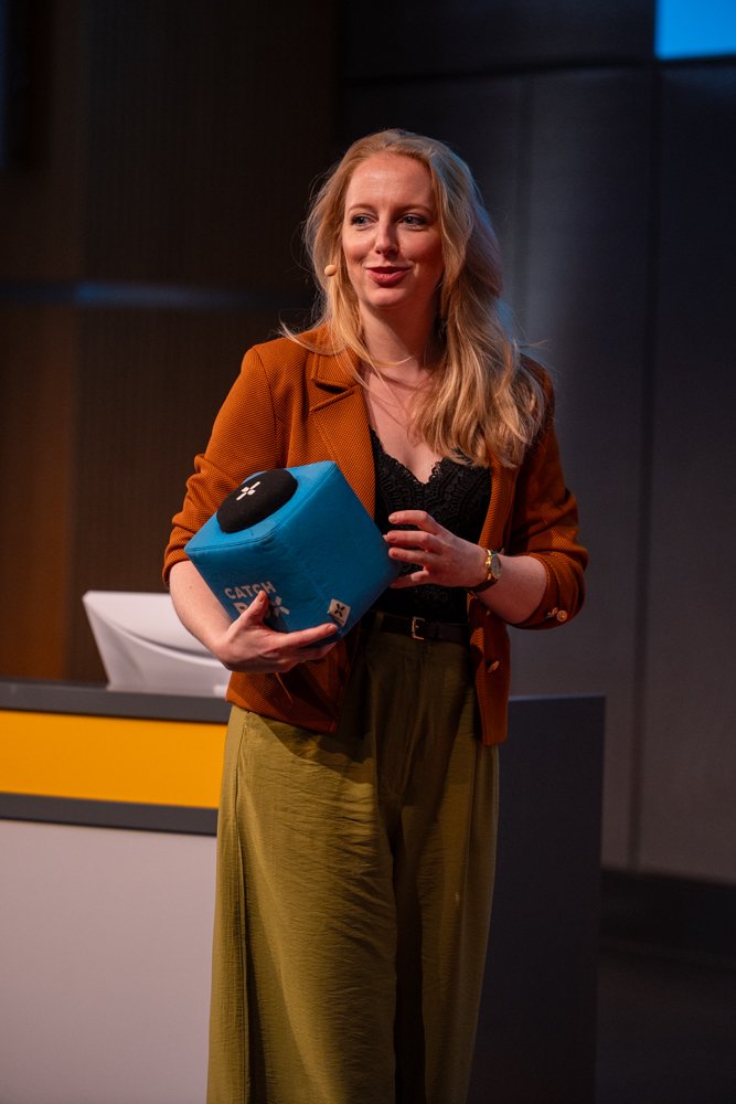 Eine Frau mit blonden Haaren, braunem Blazer, schwarzem Oberteil und einer goldenen Hose steht auf einer Bühne, hält einen blauen Gegenstand in der Hand und hat ein Mikrofon im Haar. Im Hintergrund ist ein Pult.