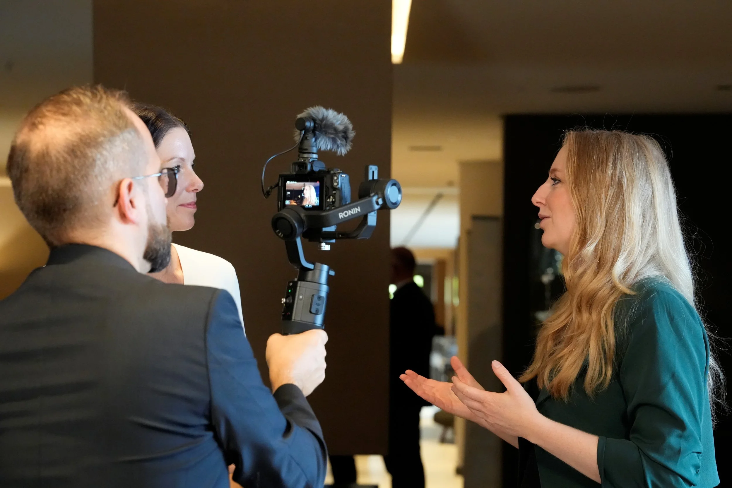 Drei Menschen im Gespräch während eines Interviews, eine Frau spricht, während ein Mann mit einer Kamera filmt, im Hintergrund sind weitere Personen in einem Innenraum.
