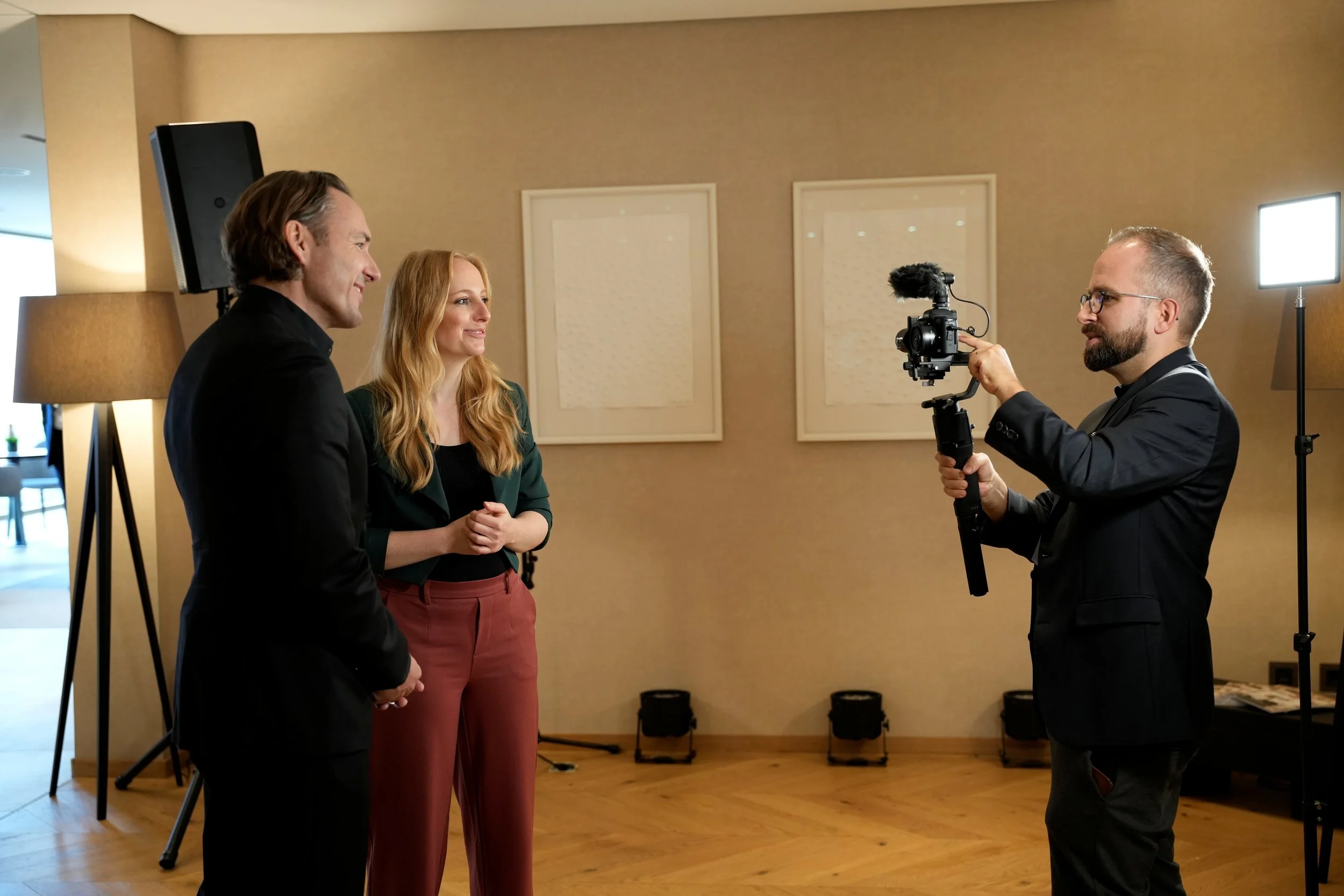 Ein Mann filmt zwei Menschen, einen Mann und eine Frau, bei einer Interviewaufnahme in einem Raum mit Holzboden und Wanddekoration.