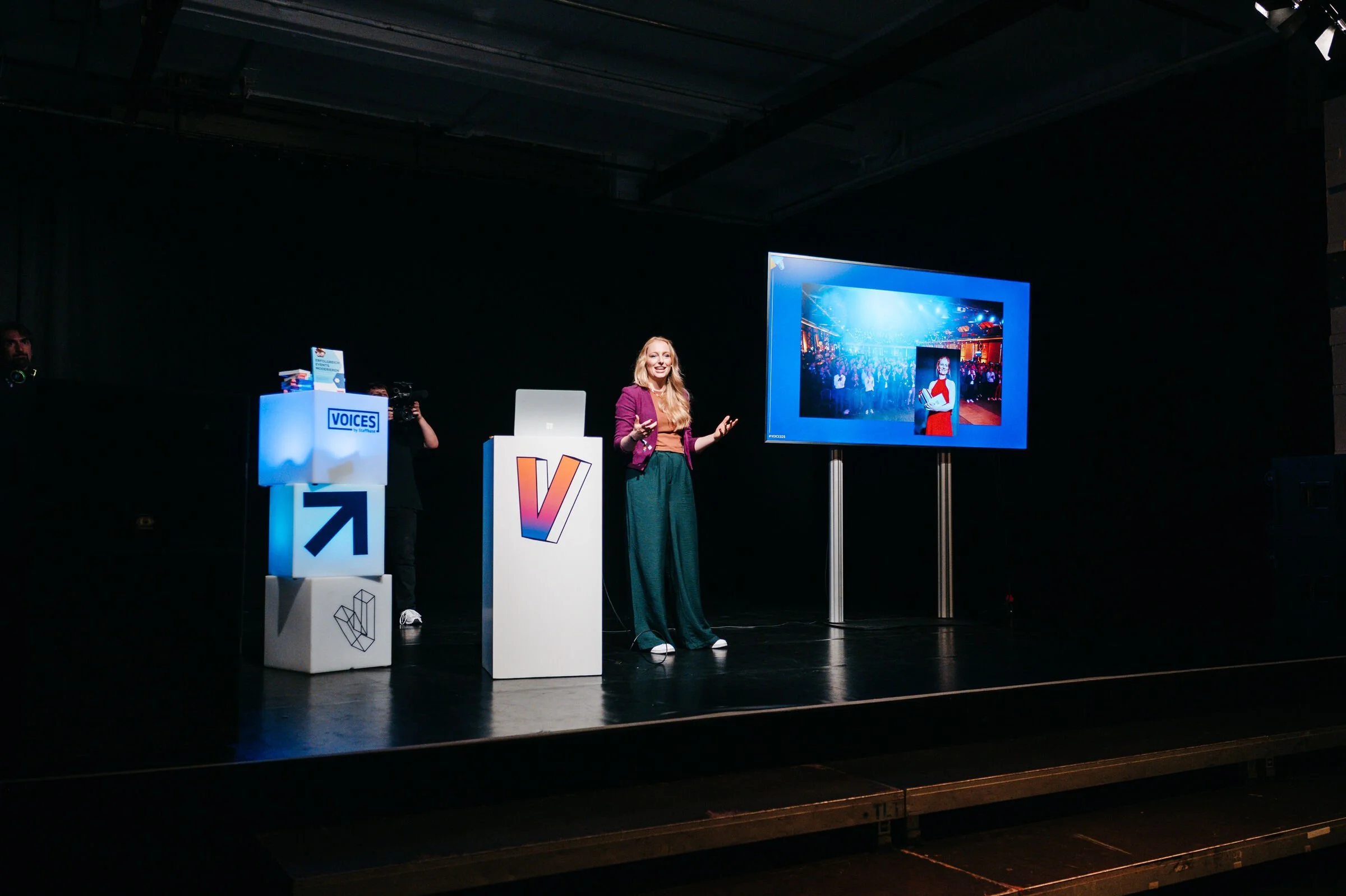 Eine Frau spricht auf einer Bühne bei einer Konferenz mit einem großen Bildschirm im Hintergrund, der eine Präsentation zeigt.