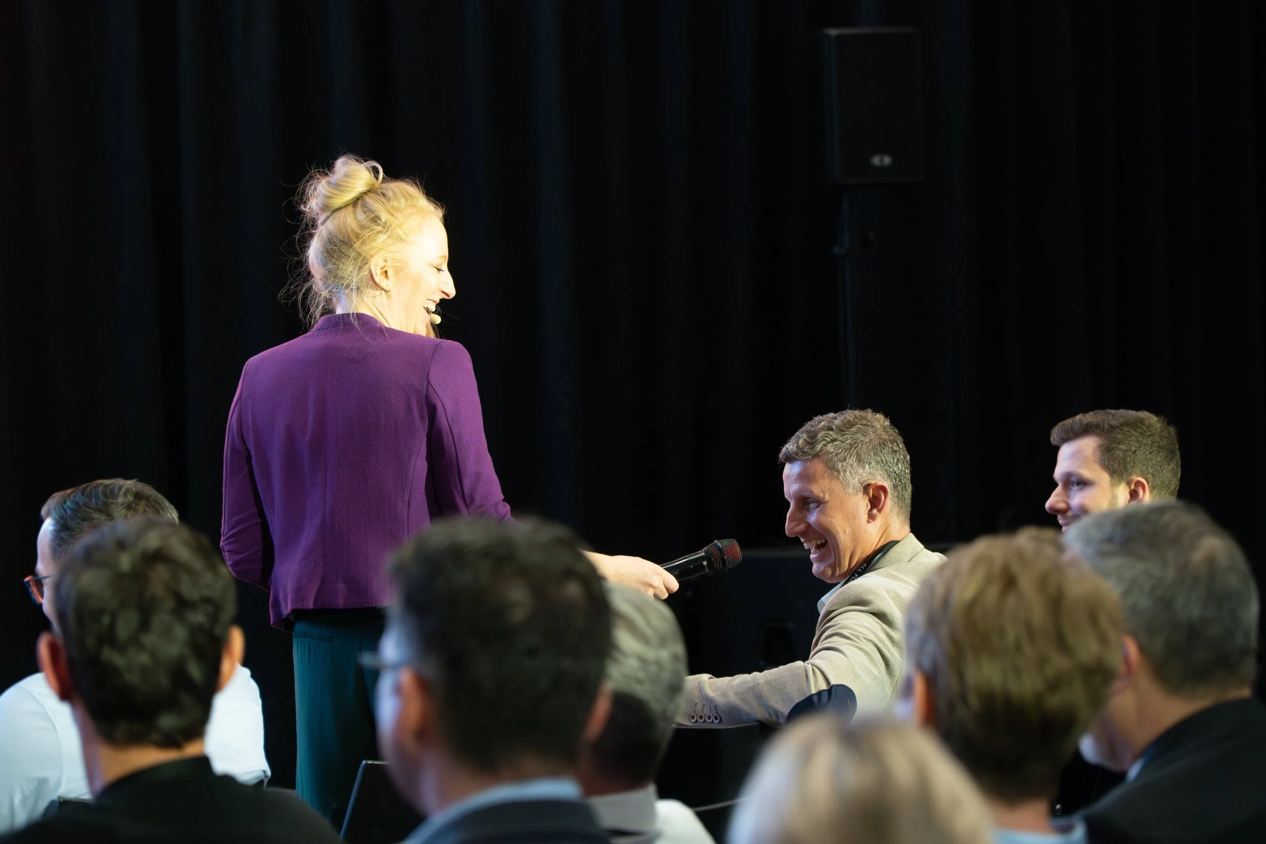 Eine Frau in lila Kleidung hält einem lachenden Mann im Publikum ein Mikrofon entgegen, während sie auf einer Bühne bei einer Konferenz oder einem Event steht. Mehrere Männer sitzen im Publikum, die Szene ist in einem dunklen Raum. 