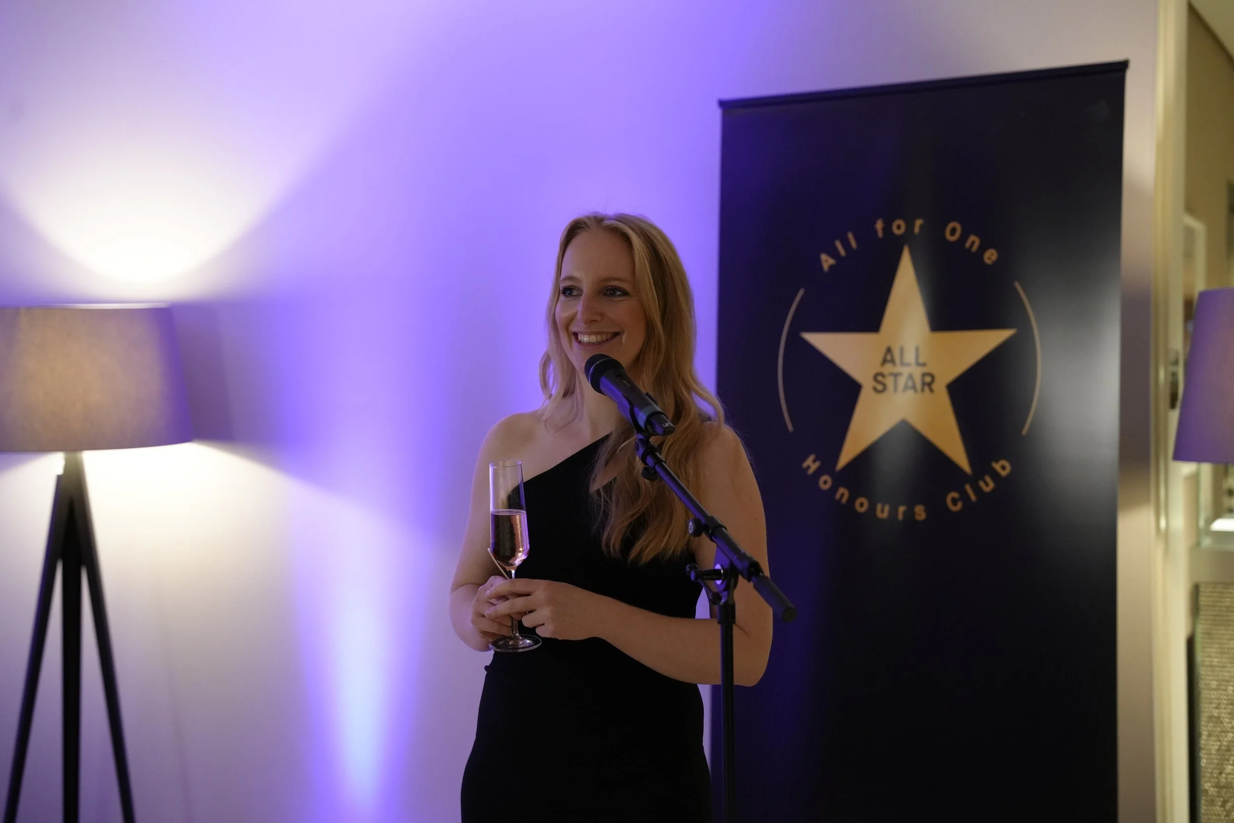 Eine lachende Frau in einem schwarzen Kleid hält ein Gläschen Sekt und spricht bei einer Veranstaltung der All Star Honours Club, im Hintergrund ein schwarzes Banner mit goldener Stern-Logo.