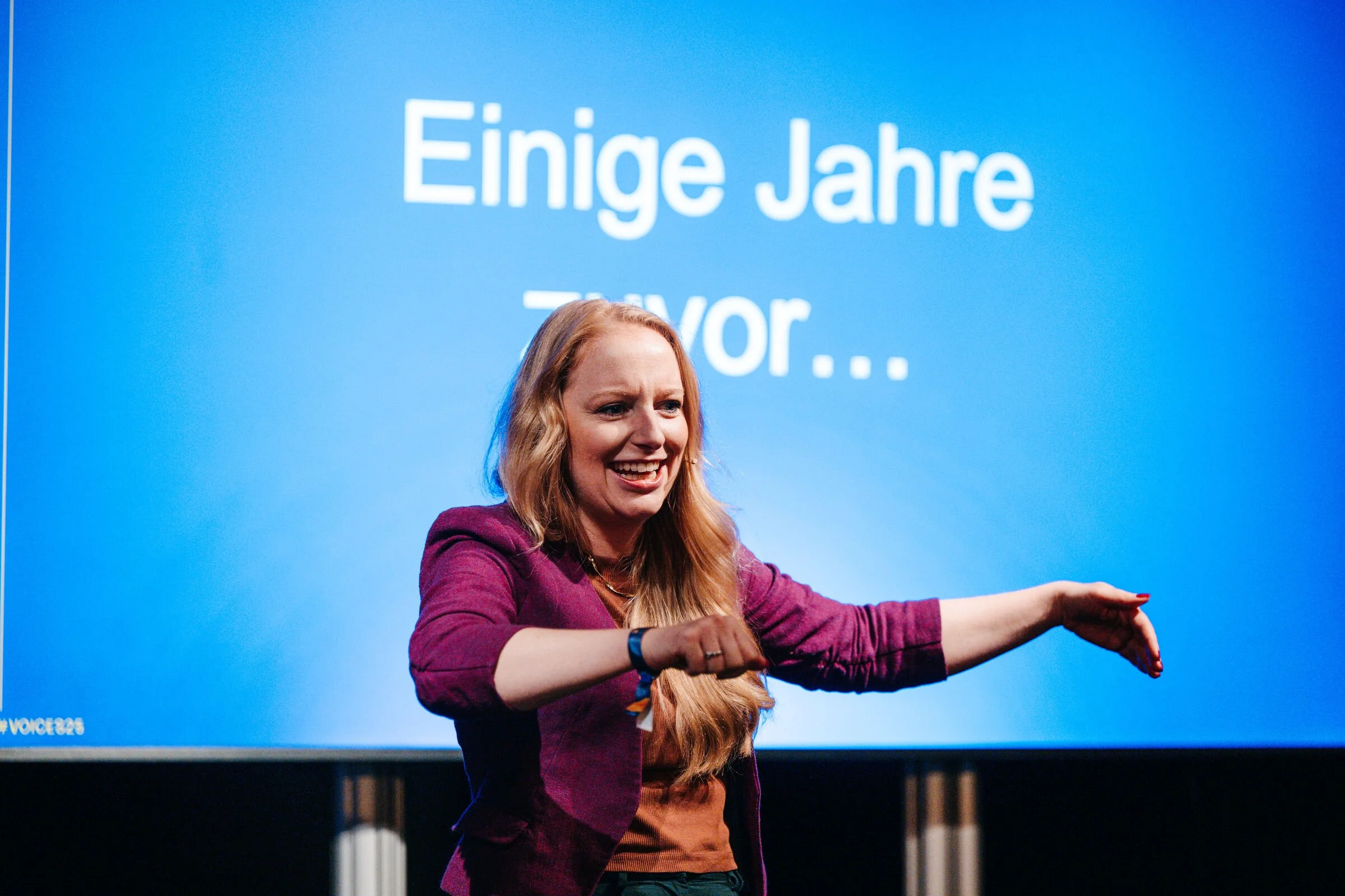 Eine Frau mit langem, blondem Haar, die auf einer Bühne steht, vor einem blauen Bildschirm mit weißen Text, lächelnd und mit einer Armbewegung.