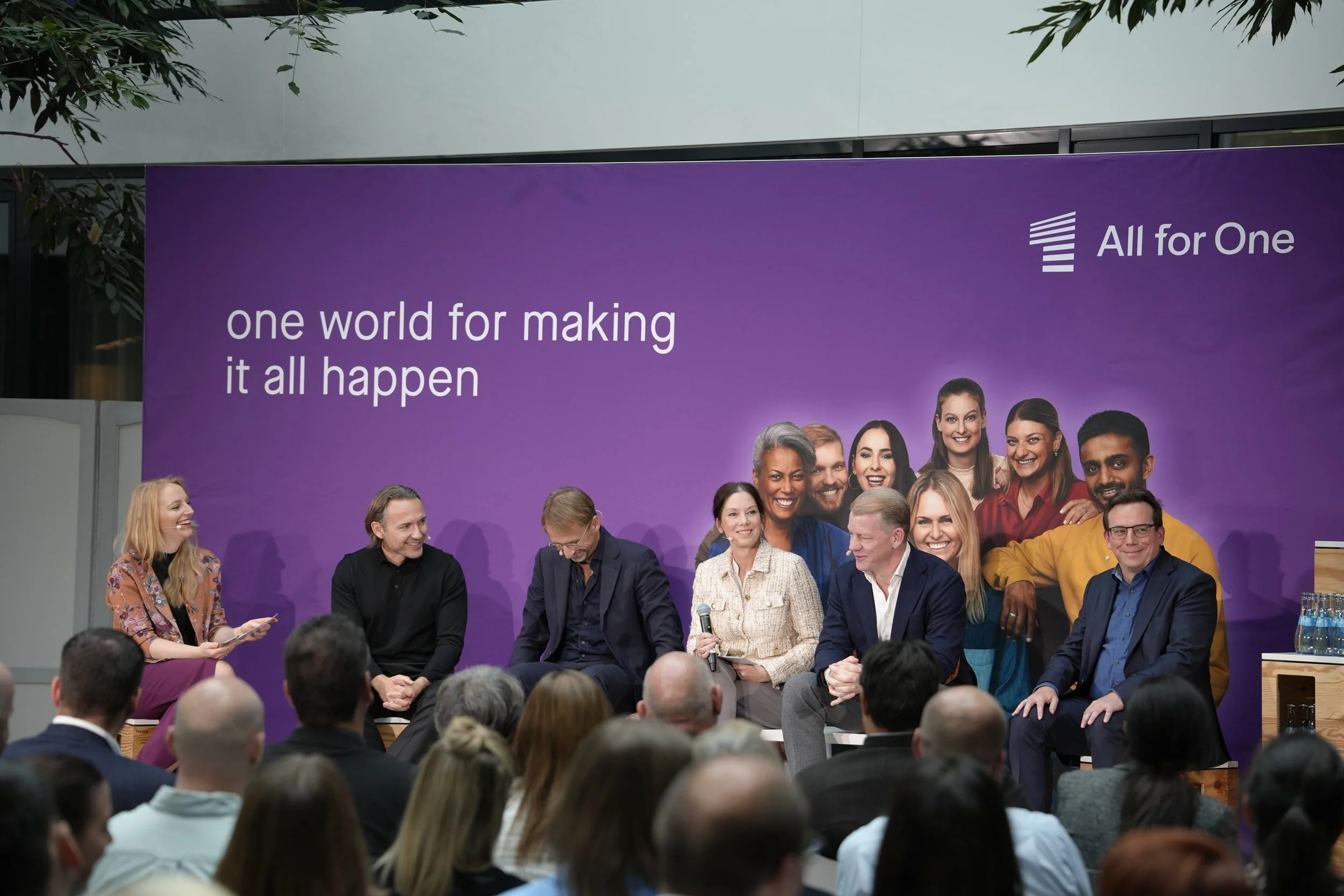 Paneldiskussion bei einer Veranstaltung mit vielen Zuschauern, im Hintergrund eine große lila Wand mit einem Logo und dem Text 'one world for making it all happen'