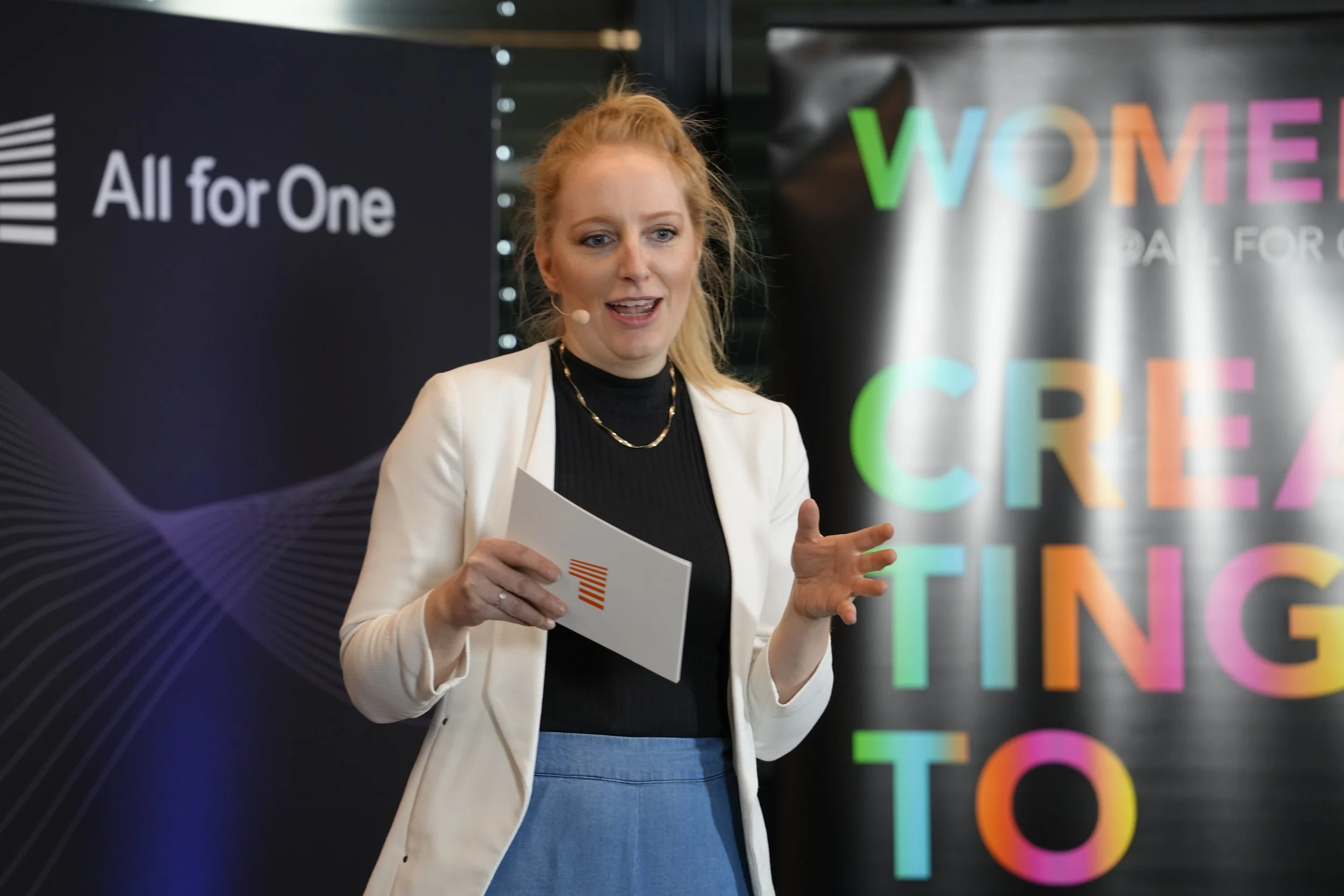 Eine Frau bei einer Präsentation, in der Nähe sind Logos und Slogans wie 'All for One' und 'Women are created to' zu sehen.
