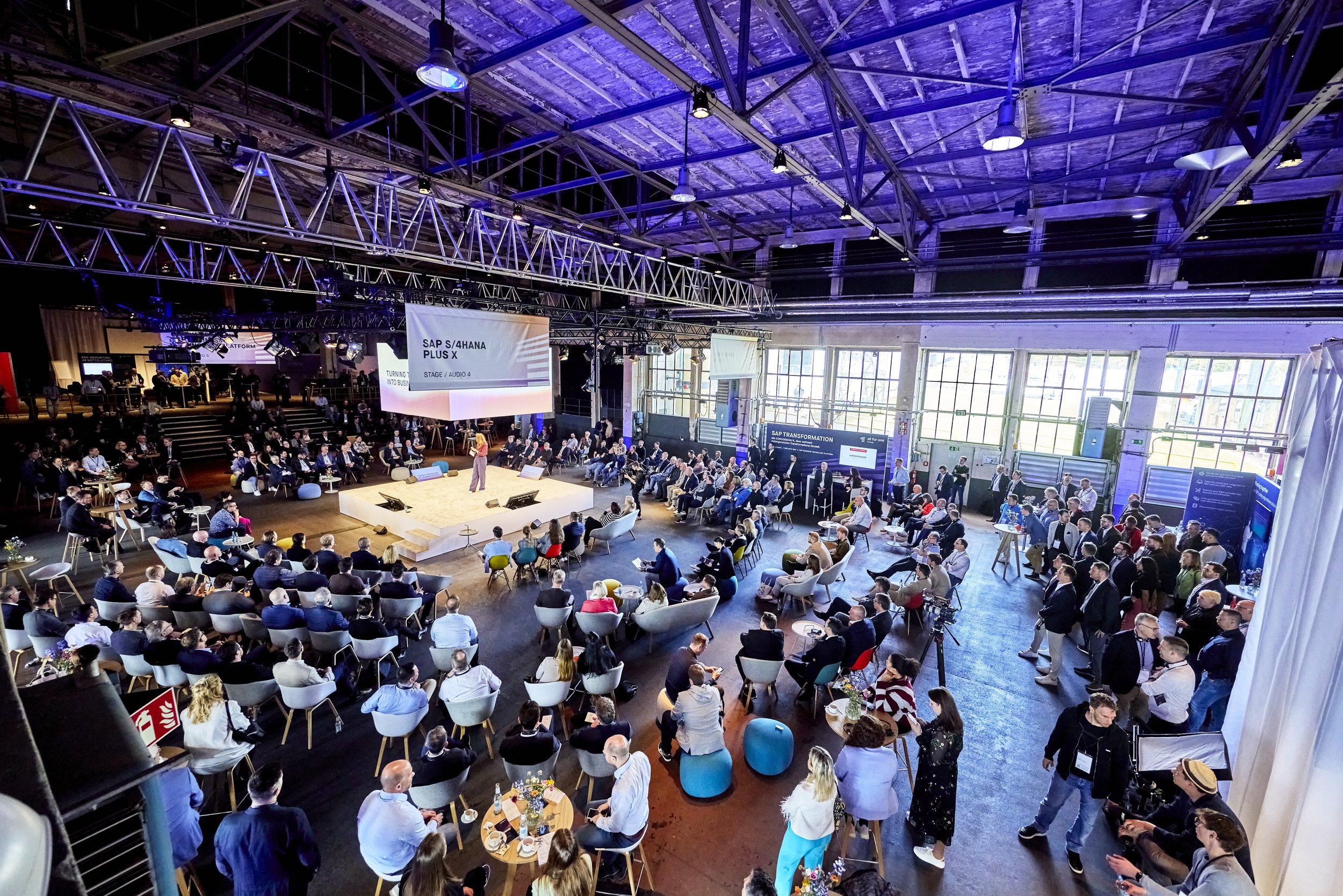Große Konferenzhalle mit vielen Teilnehmern, Live-Moderation auf der Bühne, große Bildschirme mit SAP-Themen, moderne industrialle Architektur mit hoher Decke und großen Fenstern.
