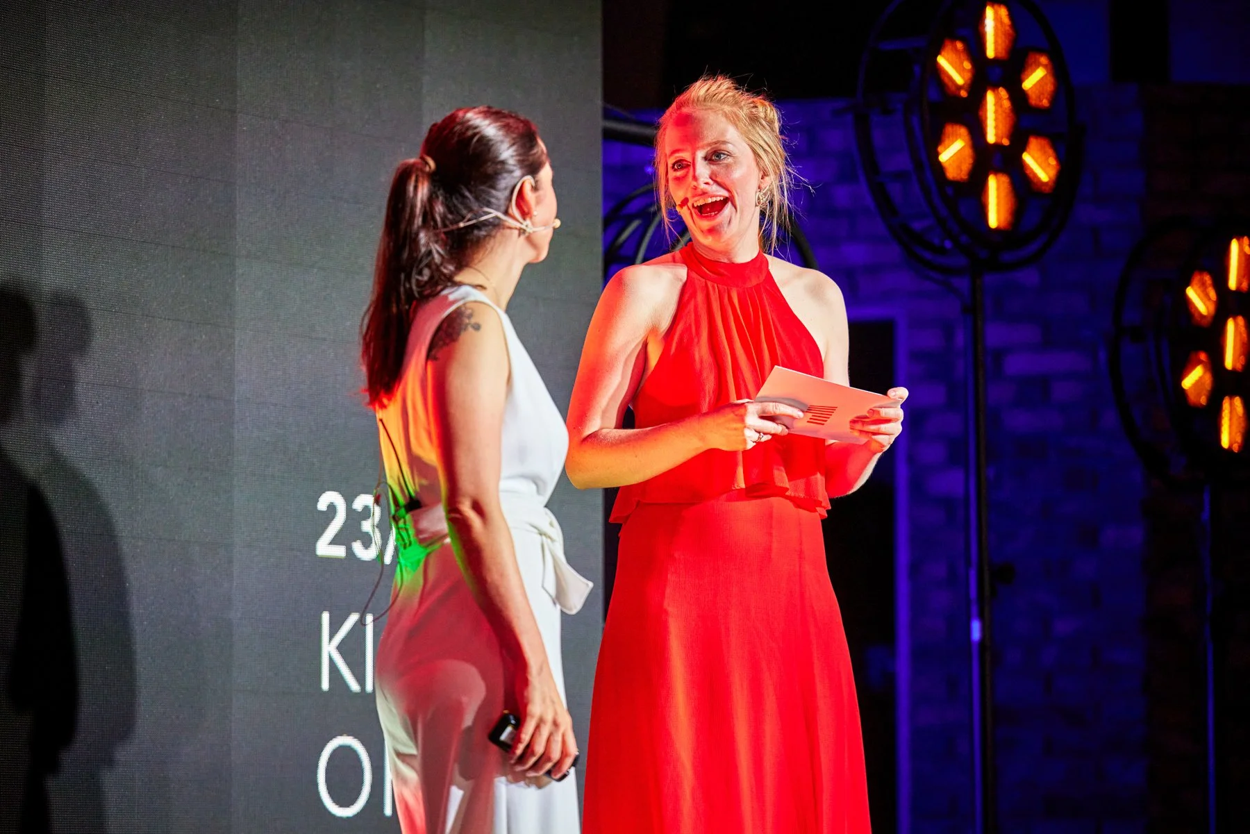 Zwei Frauen stehen auf einer Bühne im Gespräch. Eine Frau trägt ein rotes Kleid, hält Papiere, lächelt und schaut die andere Frau an. Die andere Frau trägt ein weißes Outfit und hält ein Mikrofon.