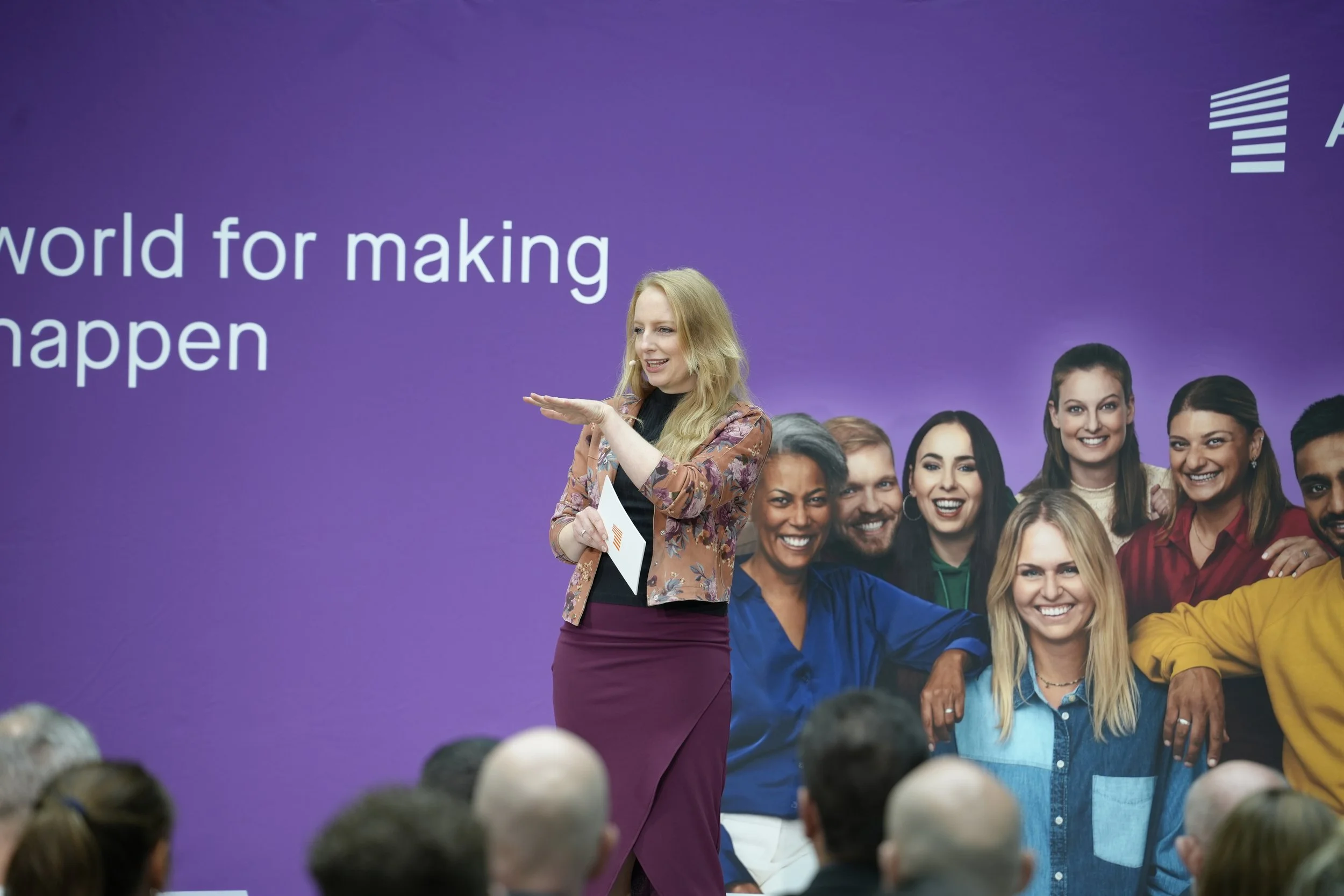Eine Frau steht vor einem Publikum bei einer Veranstaltung mit einem purplefarbigen Hintergrund mit Gruppenfoto verschiedener lachender Menschen.