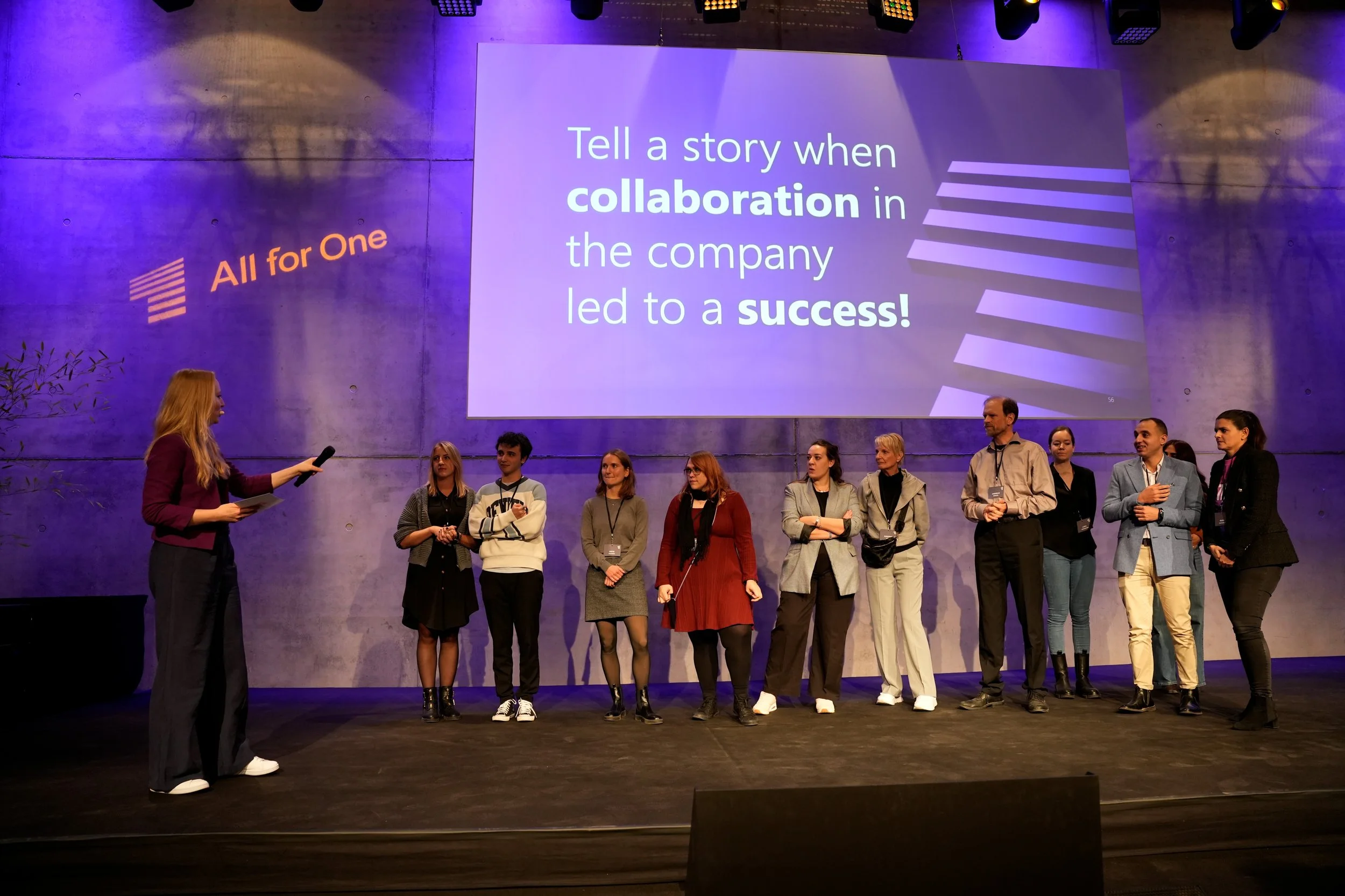 Mehrere Menschen stehen auf einer Bühne während einer Präsentation. Es gibt einen großen Bildschirm im Hintergrund mit Text, der sagt: ‚Tell a story when collaboration in the company led to a success!‘, und auf der linken Seite eine Beschriftung: ‚Al