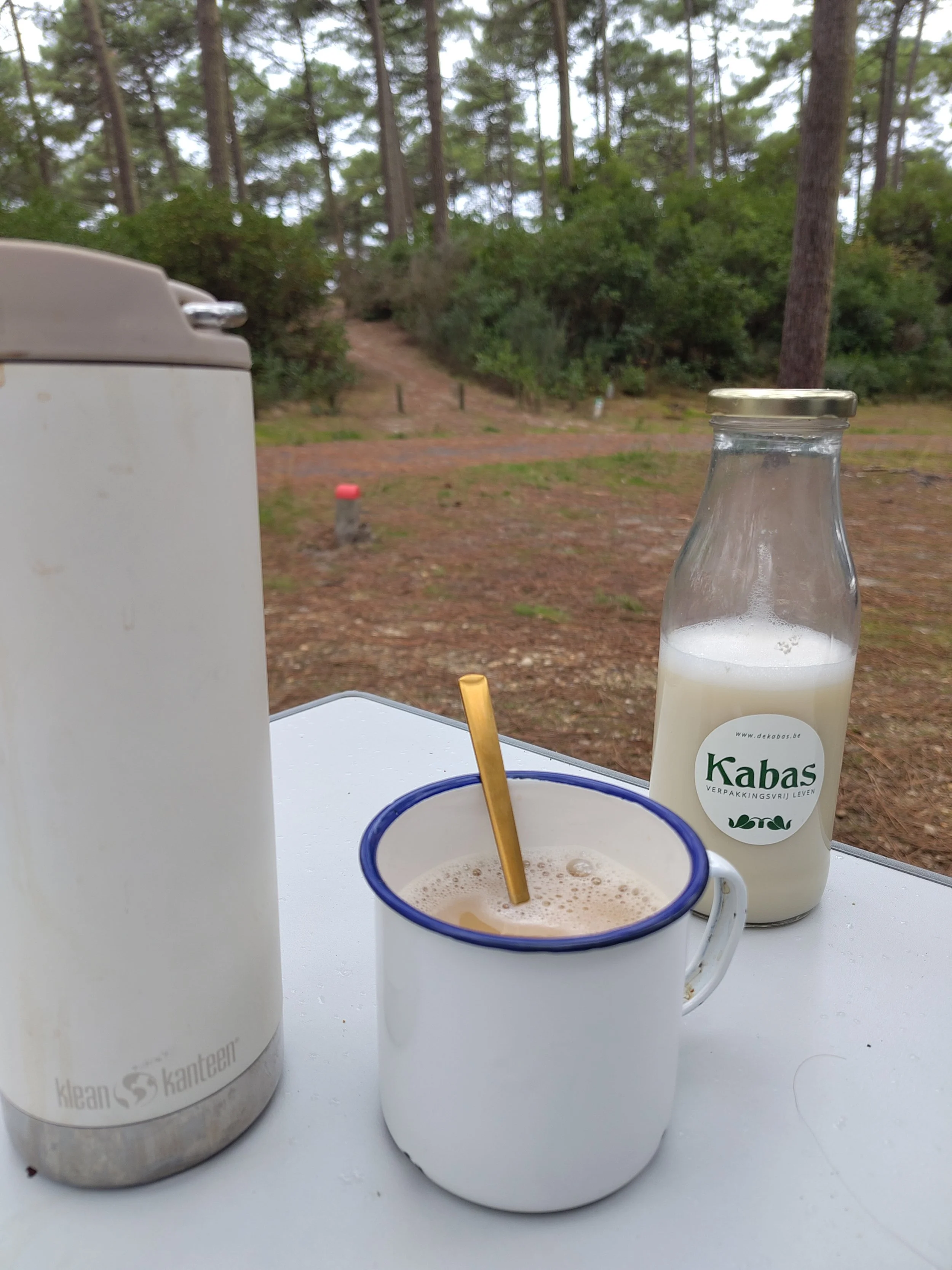 Een kopje thee met zelfgemaakte plantaardige melk, een flesje melk en een thermos van klean kanteen op een witte tafel in een bosrijke omgeving.