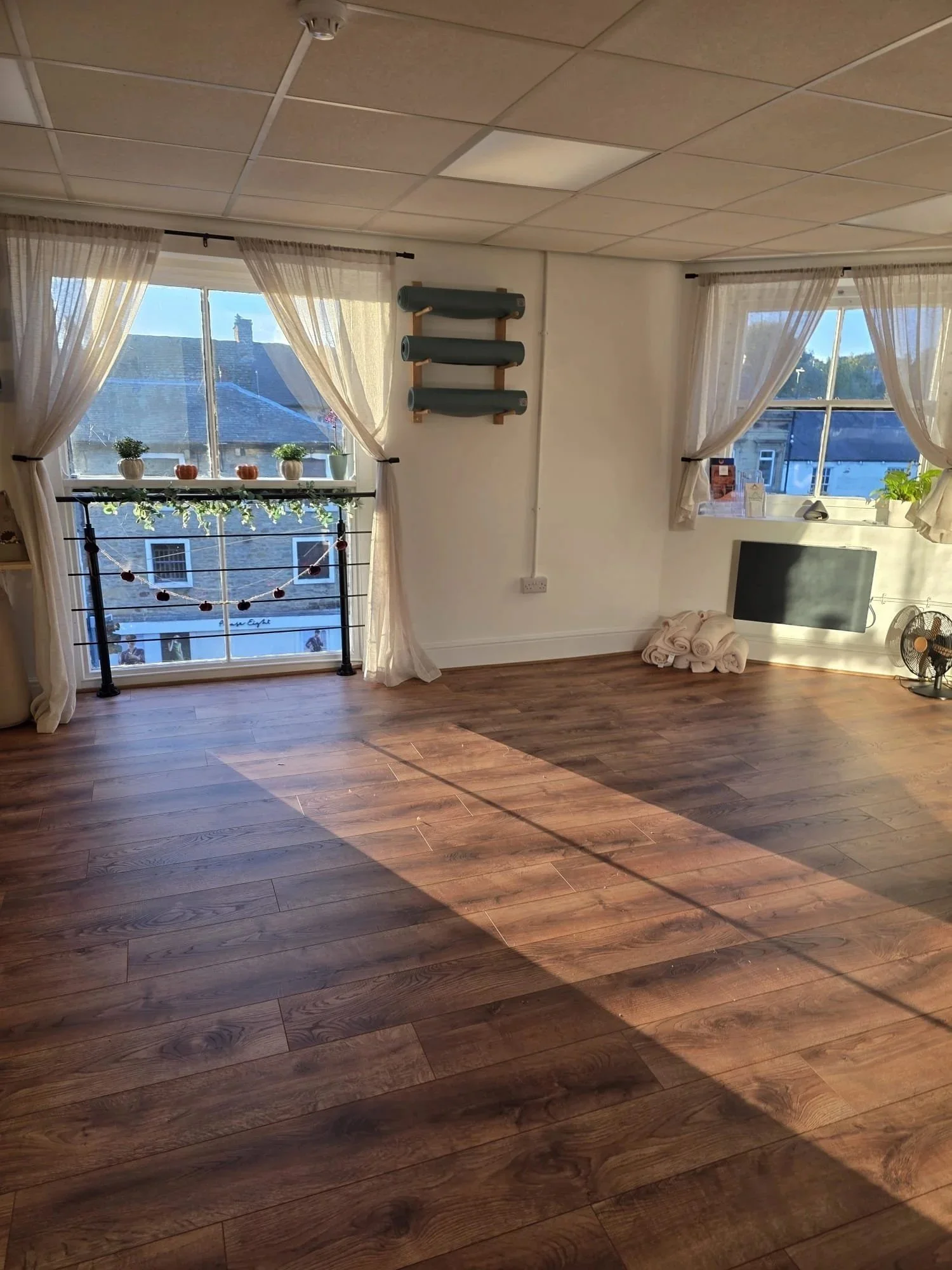 Empty room with wooden flooring, large windows with sheer curtains, potted plants on the window sills, a wall-mounted shelf with rolled mats, a small stack of towels, a computer monitor, a small fan, and sunlight streaming in.
