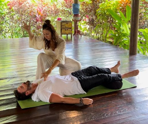 Woman performing a spiritual or healing practice over a man lying on a yoga mat outdoors surrounded by trees and plants.