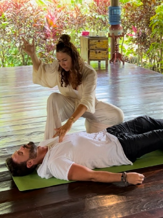 A woman practicing Kundalini Activation (KAP)healing on a man lying on a mat on a wooden floor, with lush greenery visible outside a covered outdoor space.