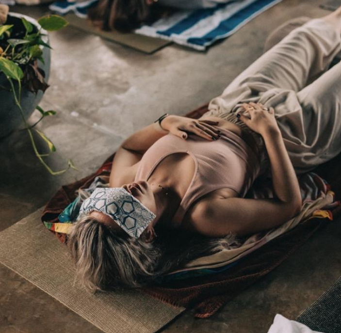 A woman lying on her side on a mat, wearing a face mask and comfortable clothing, in what appears to be a relaxed or resting state inside a room with a concrete floor.