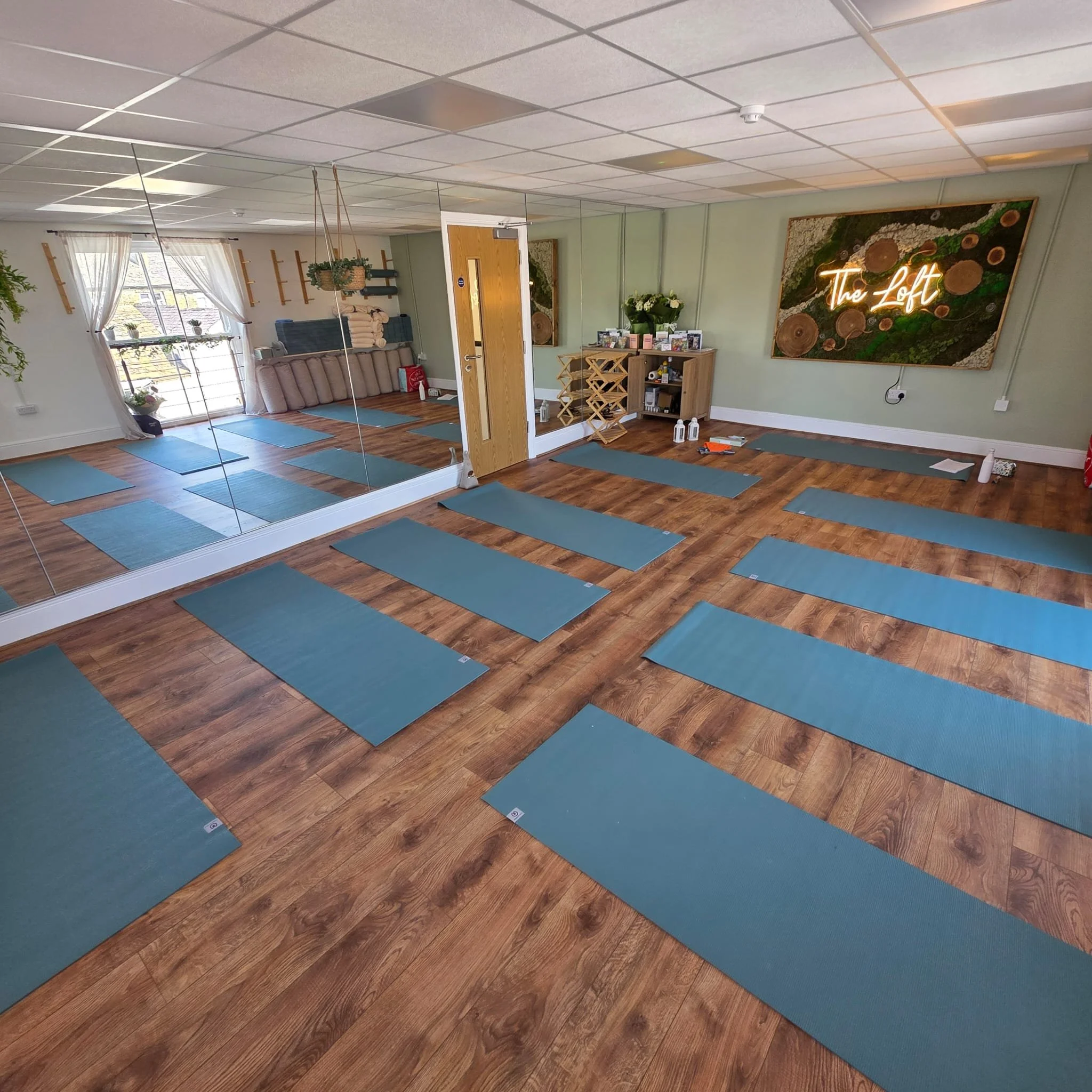 A yoga or fitness studio with blue mats arranged on a wooden floor, mirrors on one wall, a window with white curtains, and a neon sign that says 'The Loft' on the wall.