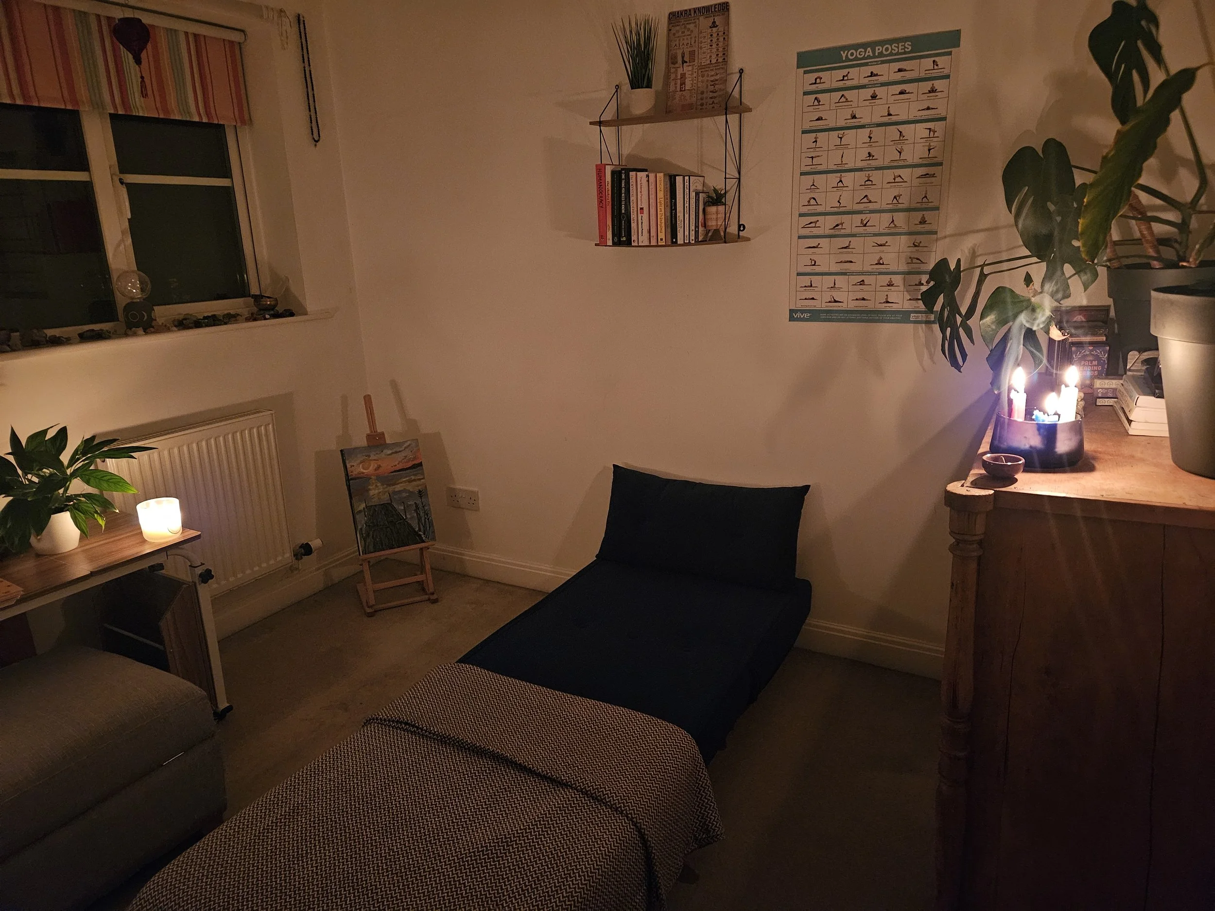 A cozy room illuminated by candles, featuring a window with a striped curtain, a radiator with a potted plant and candle on a wooden table, an easel with a landscape painting, a single bed with dark linens, a yoga poses poster on the wall, a small shelf with books and decorative items, and a chest of drawers with more candles, plants, and books.