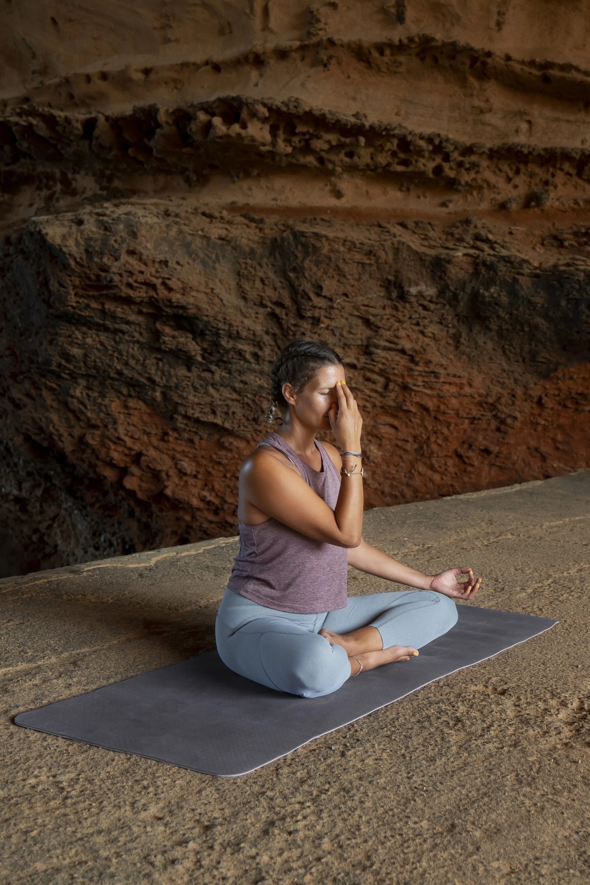 A woman practicing yoga on a mat in a rocky outdoor setting, sitting cross-legged with one hand on her knee and the other touching her face, eyes closed in meditation.