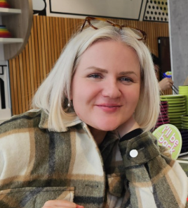 A woman with blonde hair smiling, wearing a plaid jacket, in a cafe or restaurant setting.
