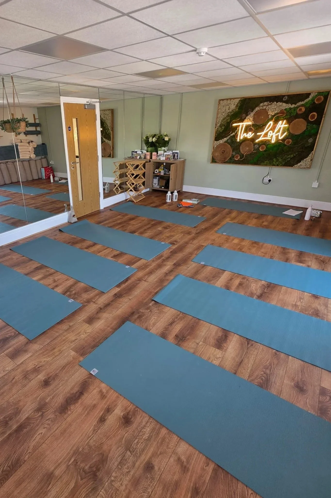 Yoga mats arranged on wooden floor in a spacious, well-lit studio with a mirror wall, decorative artwork, neon sign reading 'The Loft', and small furniture and plants in the background.