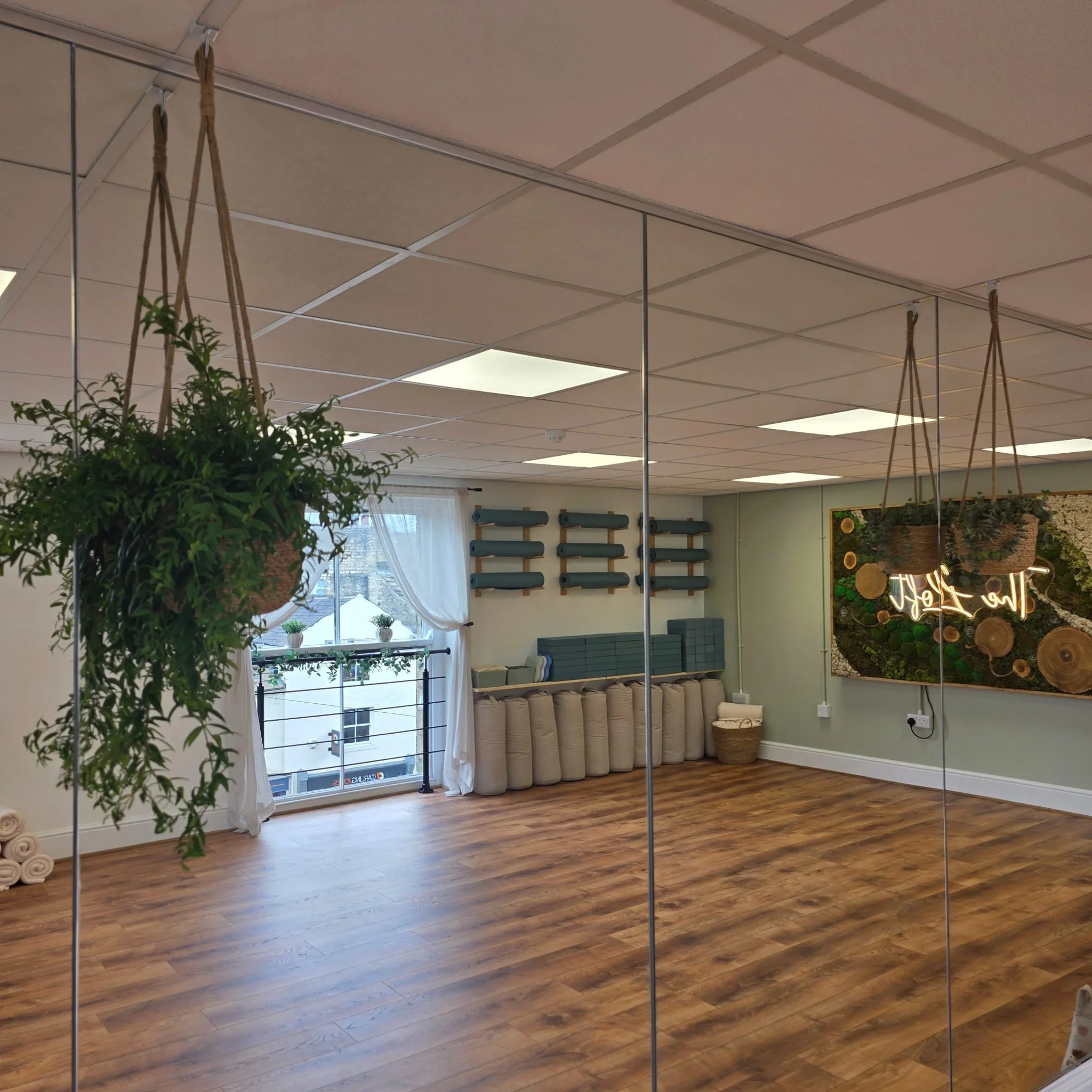 Interior of a yoga studio with hanging plants, a mirrored wall, wooden flooring, and a wall with yoga mats and decorative art.