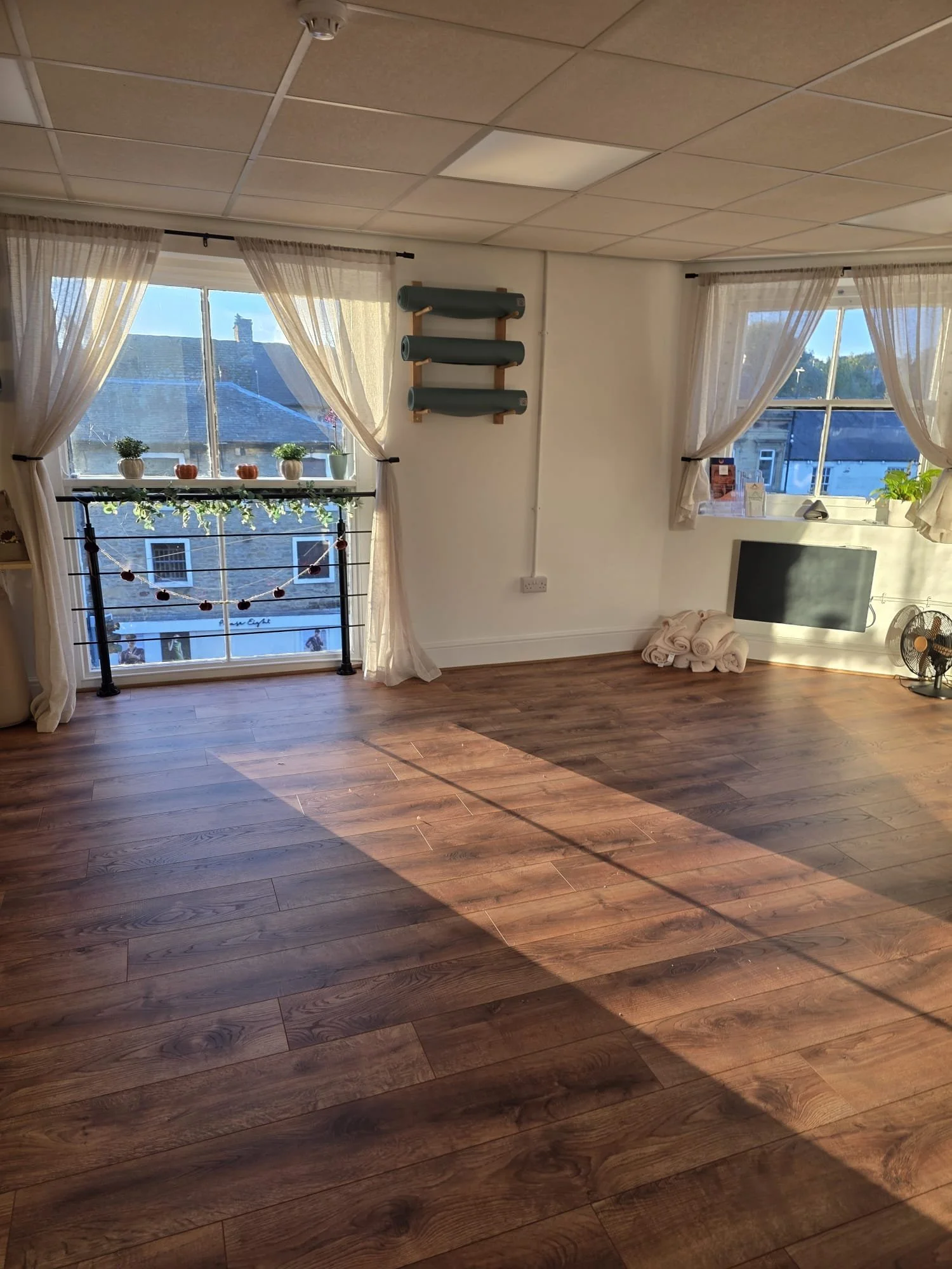 Bright, empty room with wooden flooring, two large windows with sheer curtains, small plants on the window sills, a black railing with decorative pumpkins and garland outside, and some rolled-up towels and a small fan on the floor.