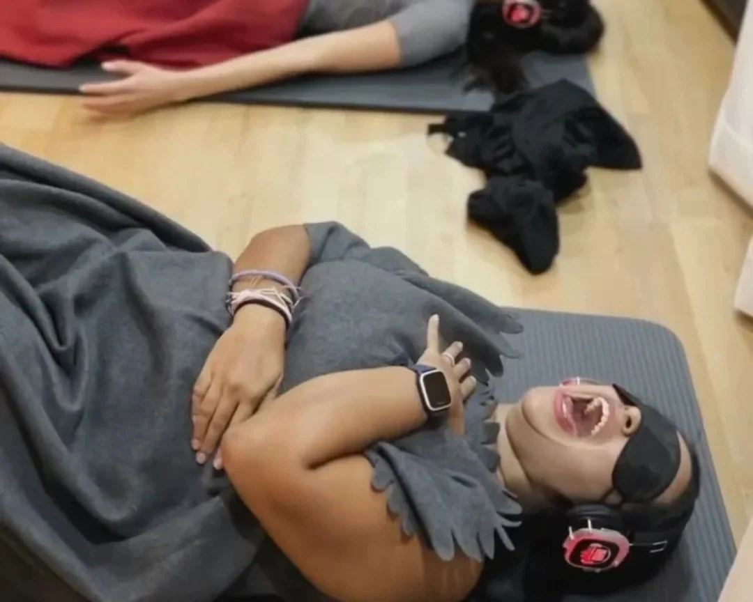 A young woman lying on her back on a yoga mat, wearing a blindfold and headphones, releasing trauma with her mouth open, arms crossed over her chest.