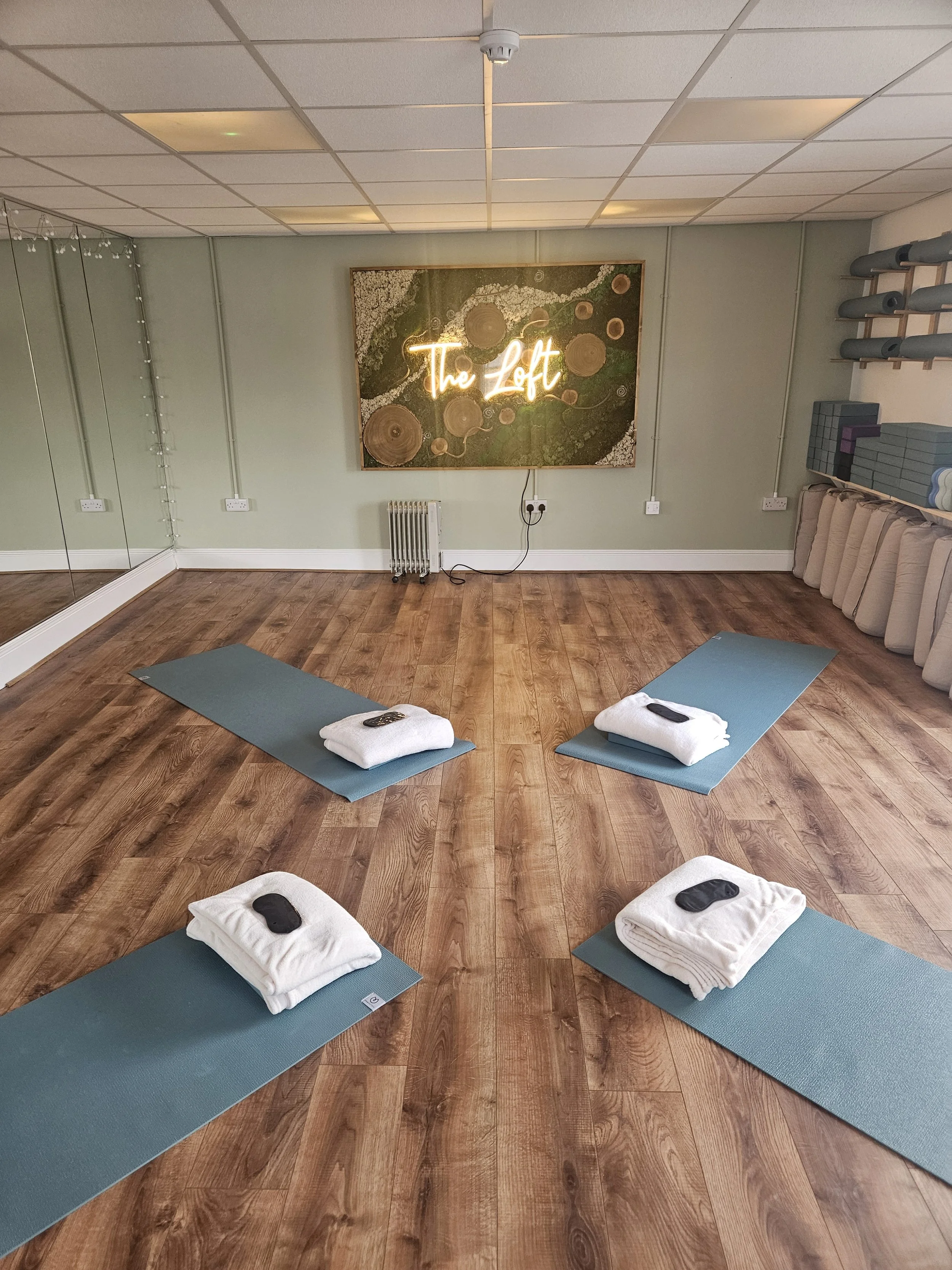 A yoga studio with four blue mats on a wooden floor, each with a folded white towel and a small black object, in front of a wall with a neon sign that reads 'The Loft' and decorative wood circles. Mirrors cover part of one wall, and shelves hold rolled-up yoga mats and blocks.