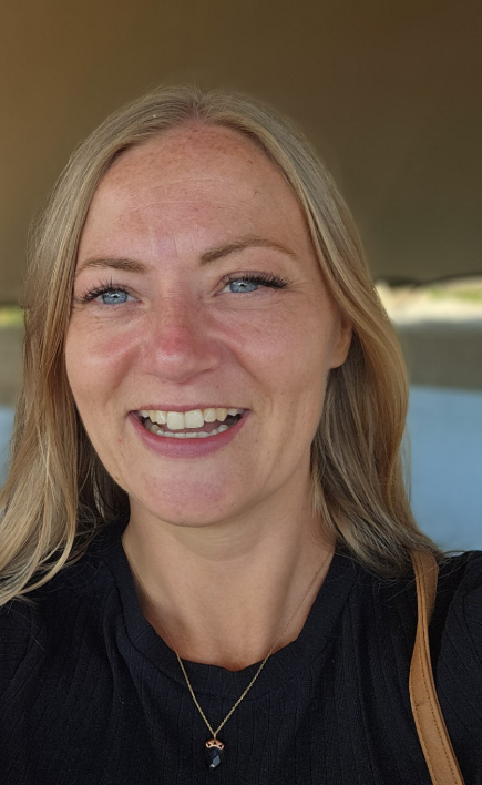 Close-up of a smiling woman, Steph Alston, with blonde hair and blue eyes wearing a black top and a necklace