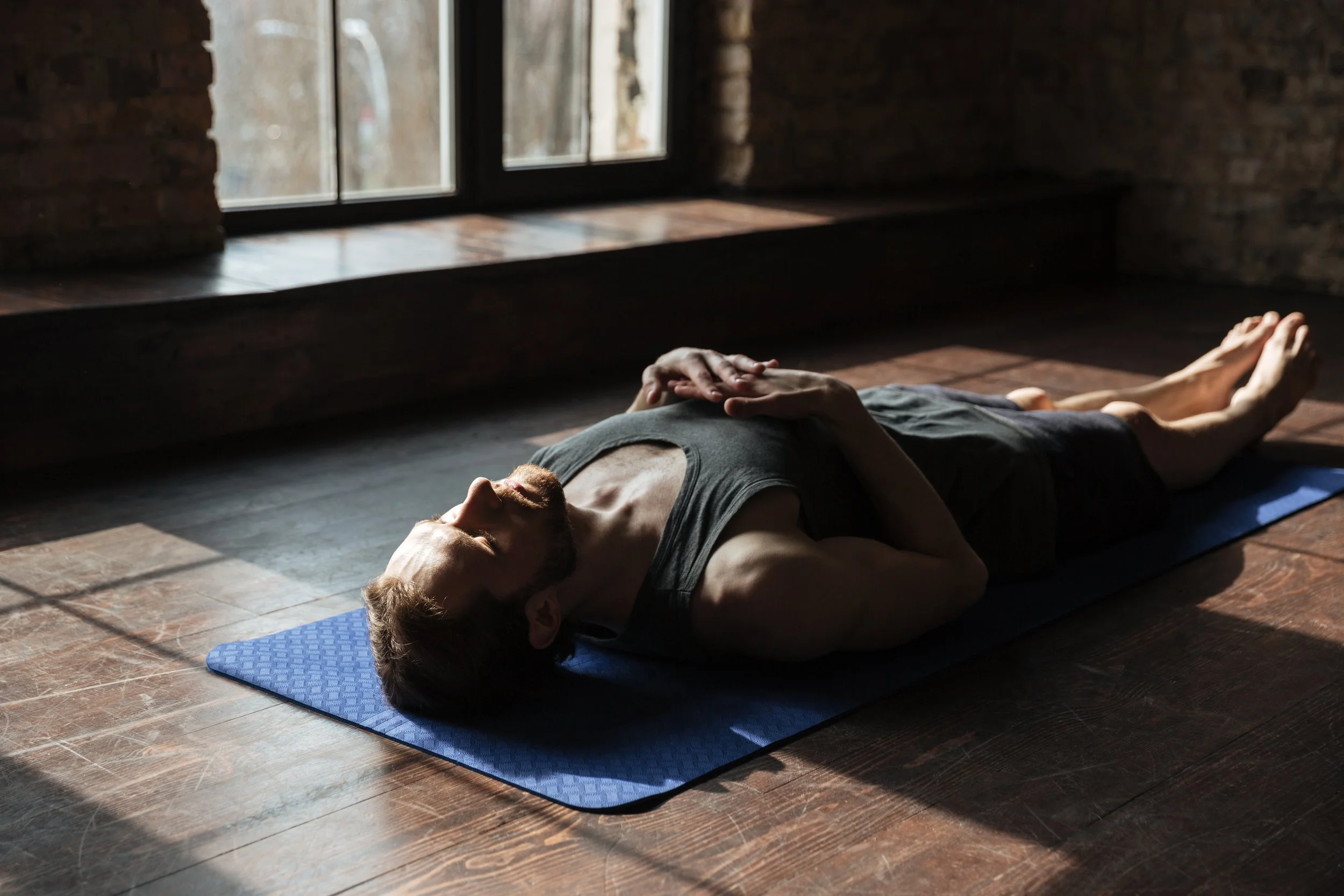 A man lying on his back on a blue yoga mat, doing a relaxation or meditation pose in a room with wooden floors and brick walls near a window.