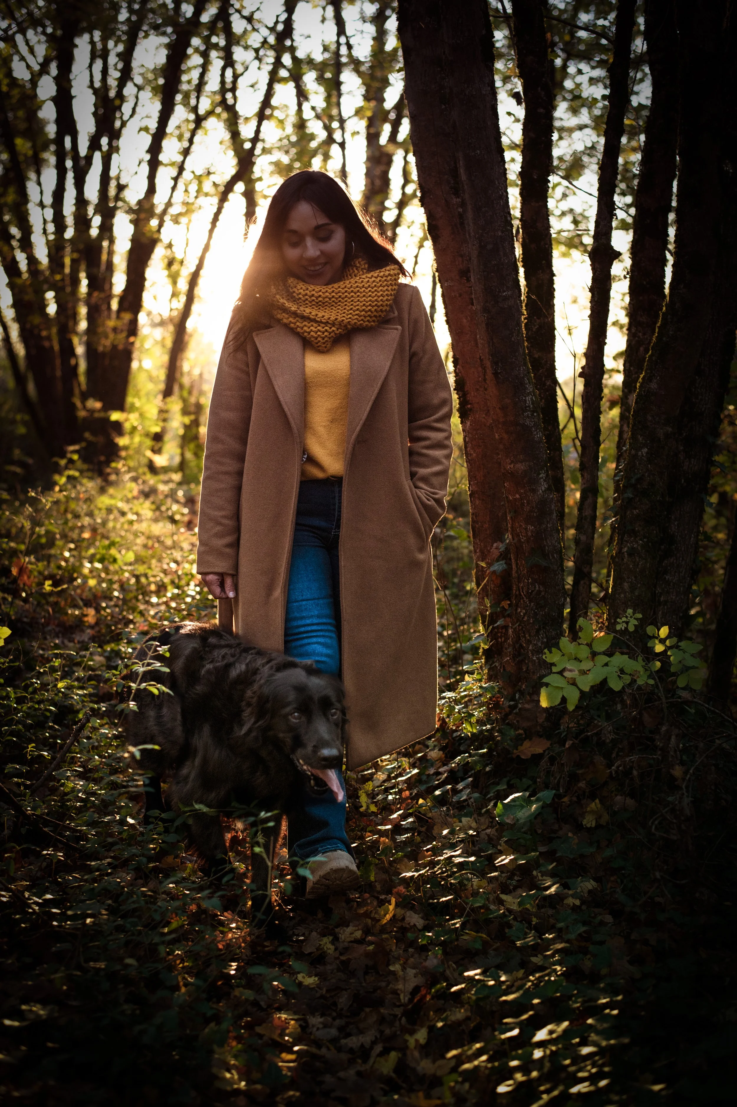 Une femme marche dans une forêt avec son chien, entourée d'arbres, lors d'un coucher de soleil, portant un manteau beige, un pull jaune et une écharpe marron, le chien est noir et marche à ses côtés.