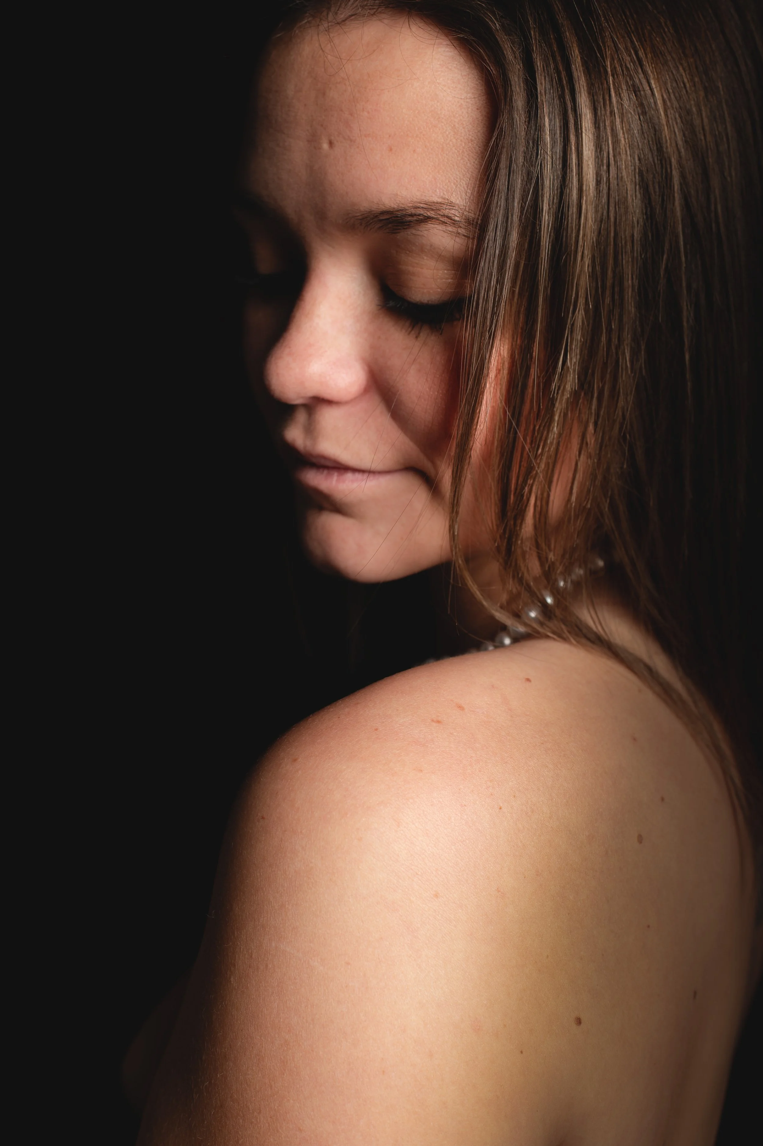 Vue rapprochée du visage et de l'épaule d'une femme avec des cheveux bruns, peau claire, yeux fermés, en contre-jour, sur fond noir, photographe royan, photographe saujon, saujon, photographe de portrait, portrait de femme, shooting