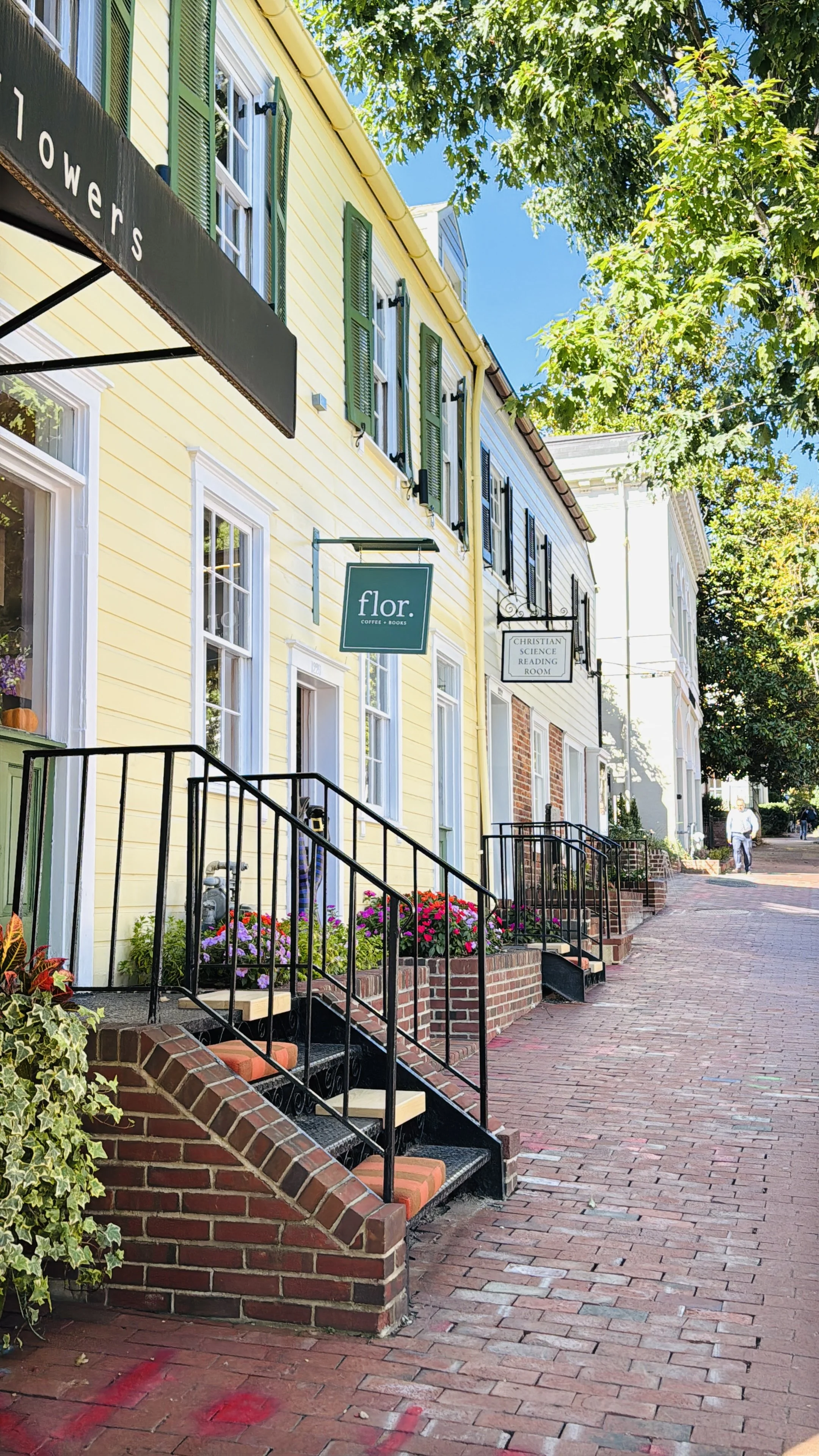A row of colorful storefronts on a brick sidewalk, with flowers in window boxes and signs for coffee and books, and a person walking in the distance.
