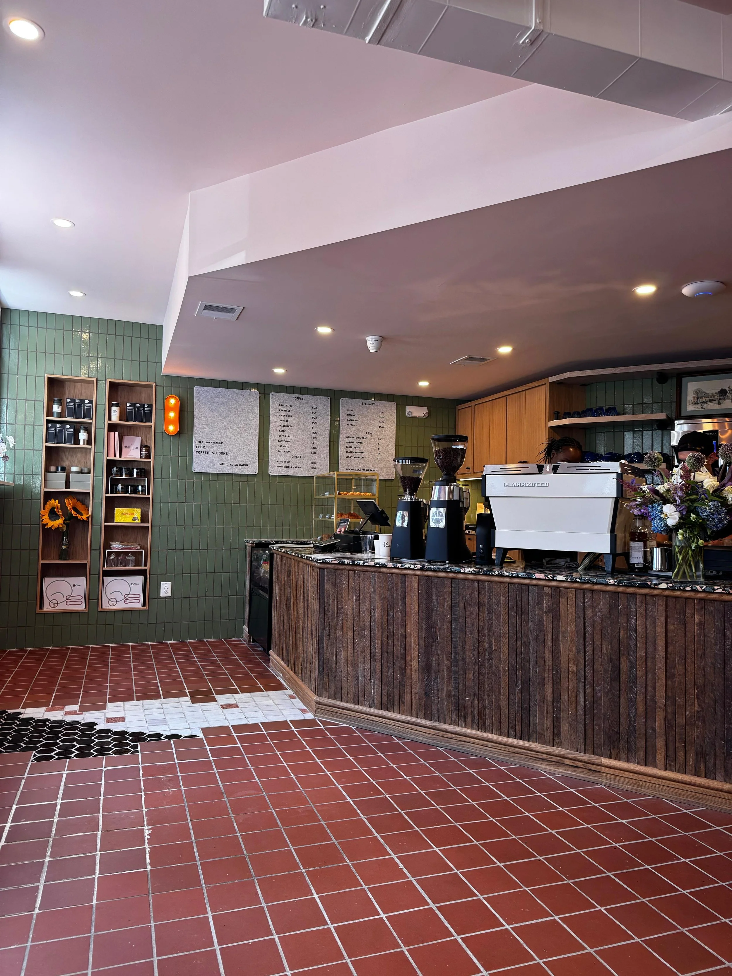 Interior of a coffee shop with a counter filled with coffee-making equipment, green tiled walls, mounted menu boards, and shelves with products. Red tiled flooring with some black and white accent tiles.