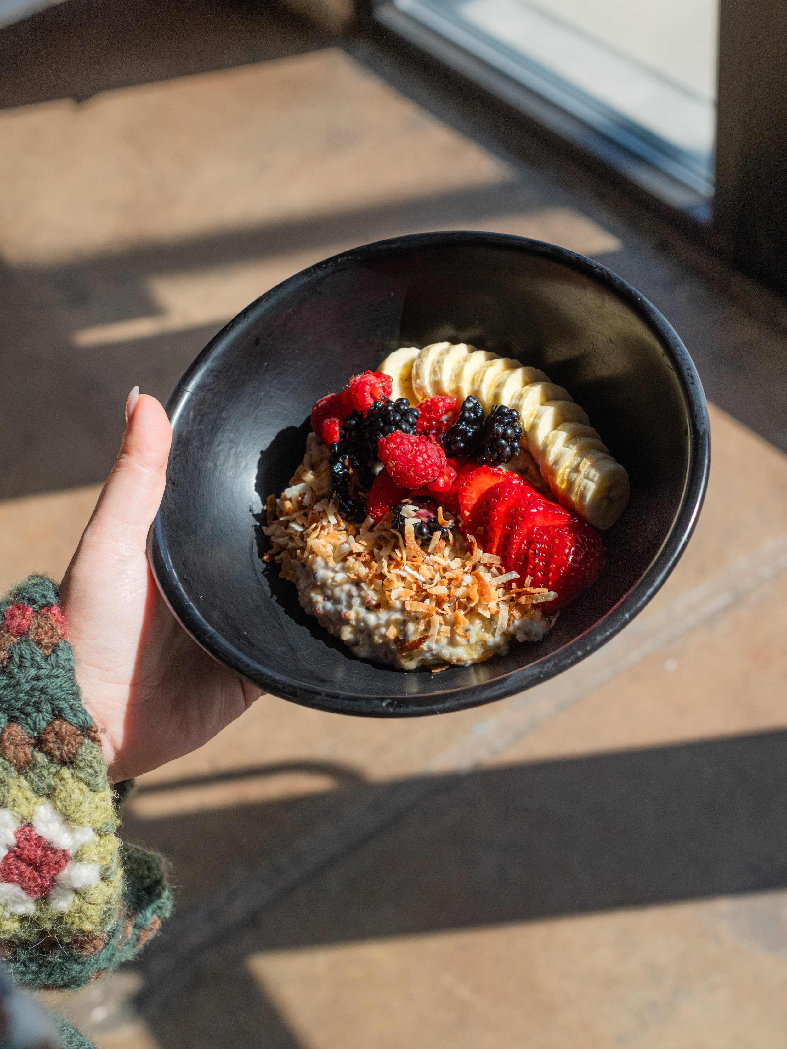 A person holding a black bowl with sliced bananas, strawberries, blackberries, raspberries, shredded coconut, and granola, with sunlight streaming in.