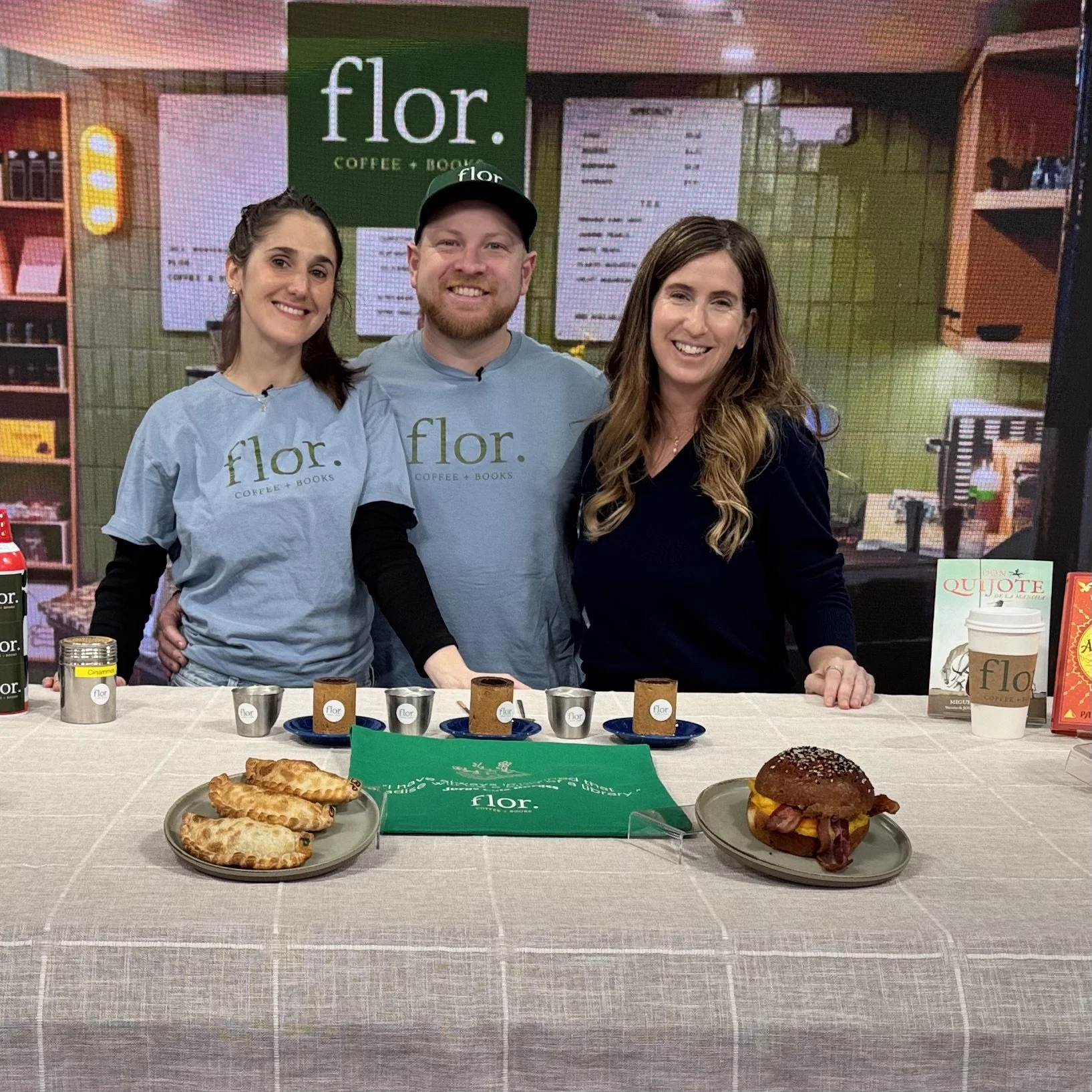Three people standing behind a table with various food items at a café. The person on the left is a woman with dark hair, smiling and wearing a light blue shirt with the word 'flor.' on it. The person in the middle is a man with a beard, wearing a light blue shirt and a black cap, also smiling. The person on the right is a woman with long wavy hair, smiling and wearing a dark top. On the table in front of them are pastries and sandwiches, and in the background, there is a green sign that reads 'flor. COFFEE + BOOKS' and cafe menu boards.