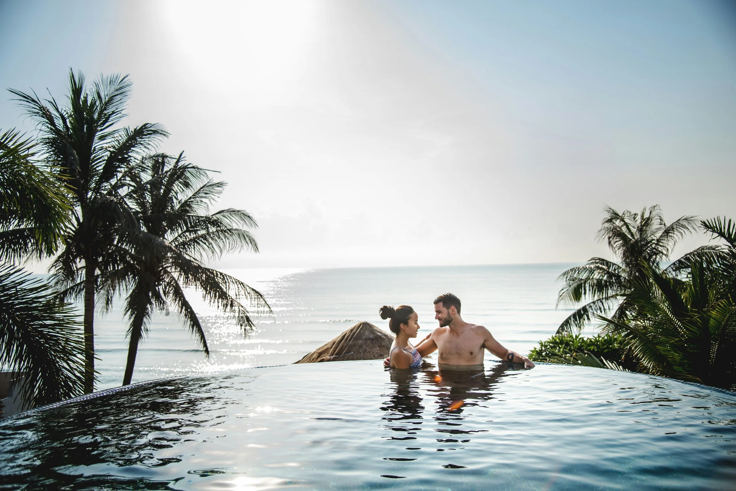 A couple in a swimming pool at the beach, surrounded by palm trees with the ocean in the background, smiling and enjoying each other's company.