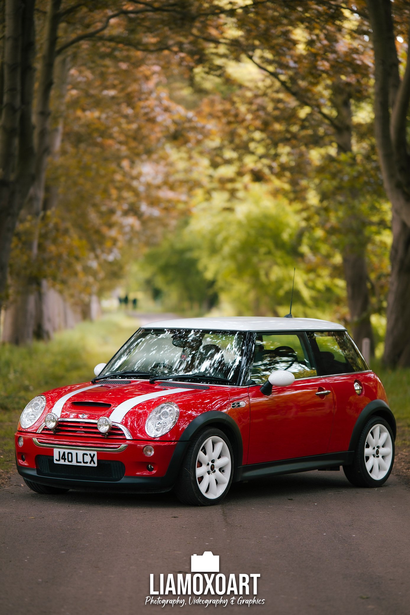 Red Mini Cooper car with white racing stripes parked on a road, with a background of trees with autumn foliage.