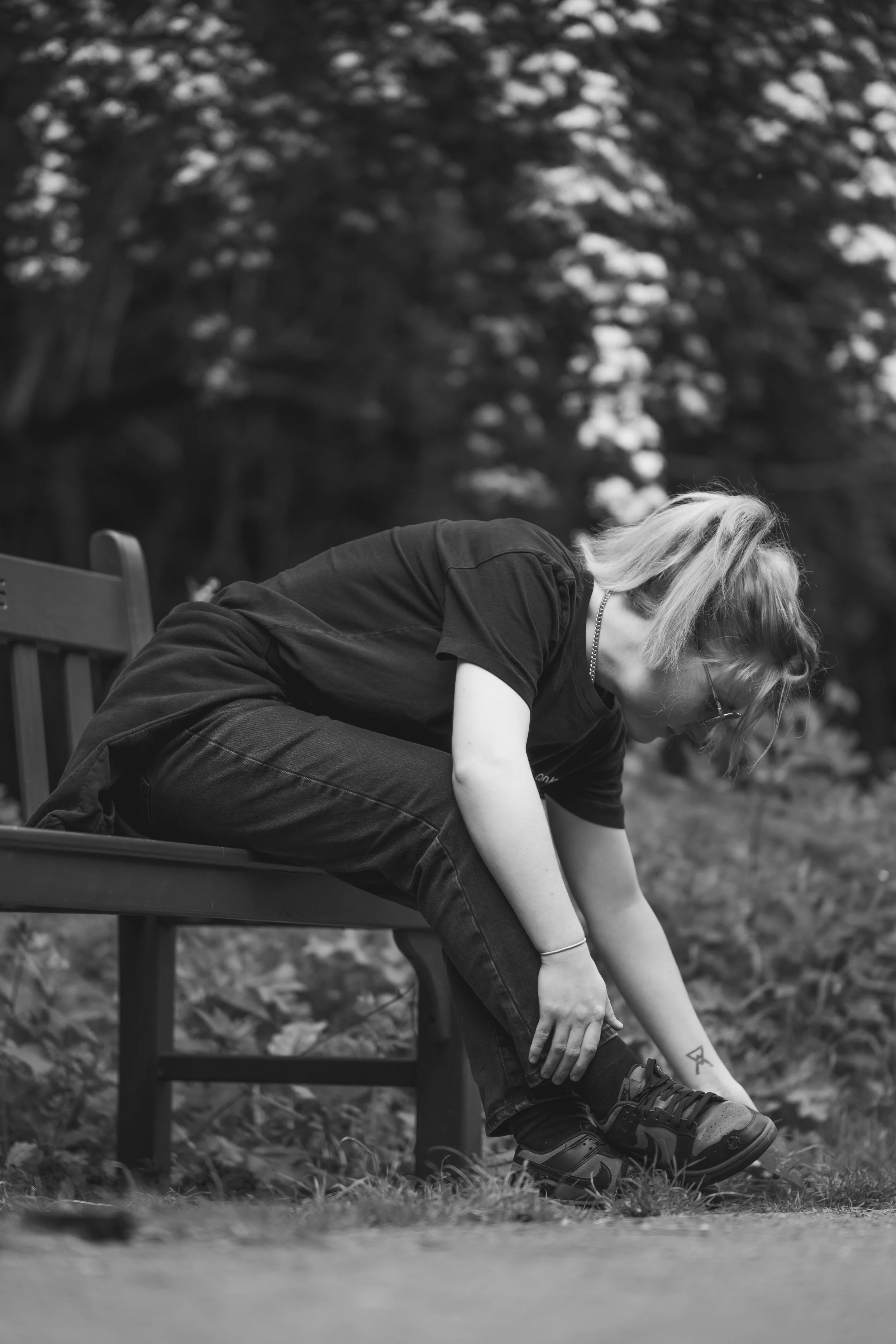 A person with light-colored hair, wearing glasses, a black t-shirt, black jeans, and sneakers, is sitting on a park bench and tying their shoelace outdoors. The photo is in black and white.