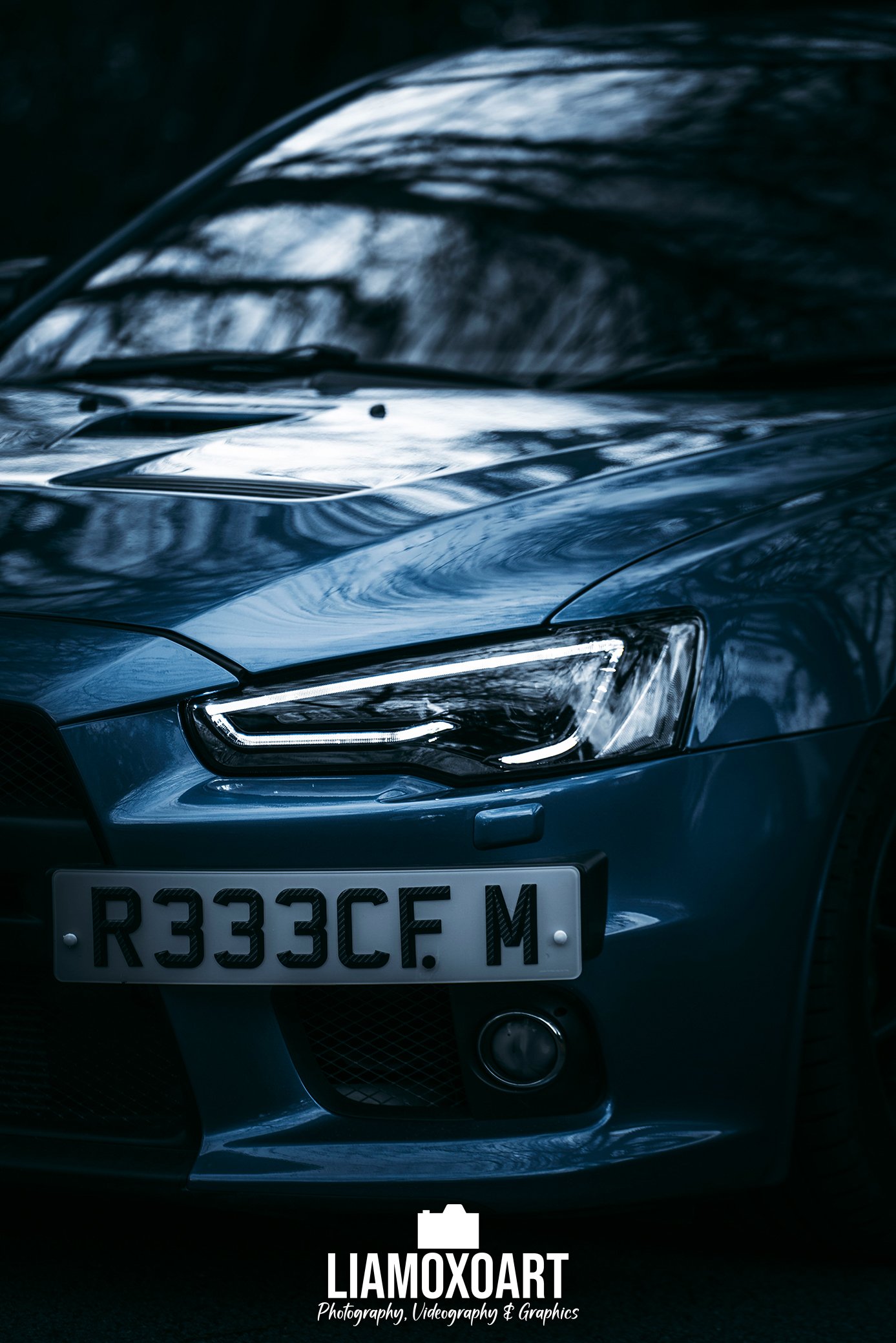 Close-up of the front part of a dark gray or black car, showing the headlight, front grille, and part of the hood. The car has a white license plate with black characters, reading R333CF M. The background is dark, with faint reflections of trees and 