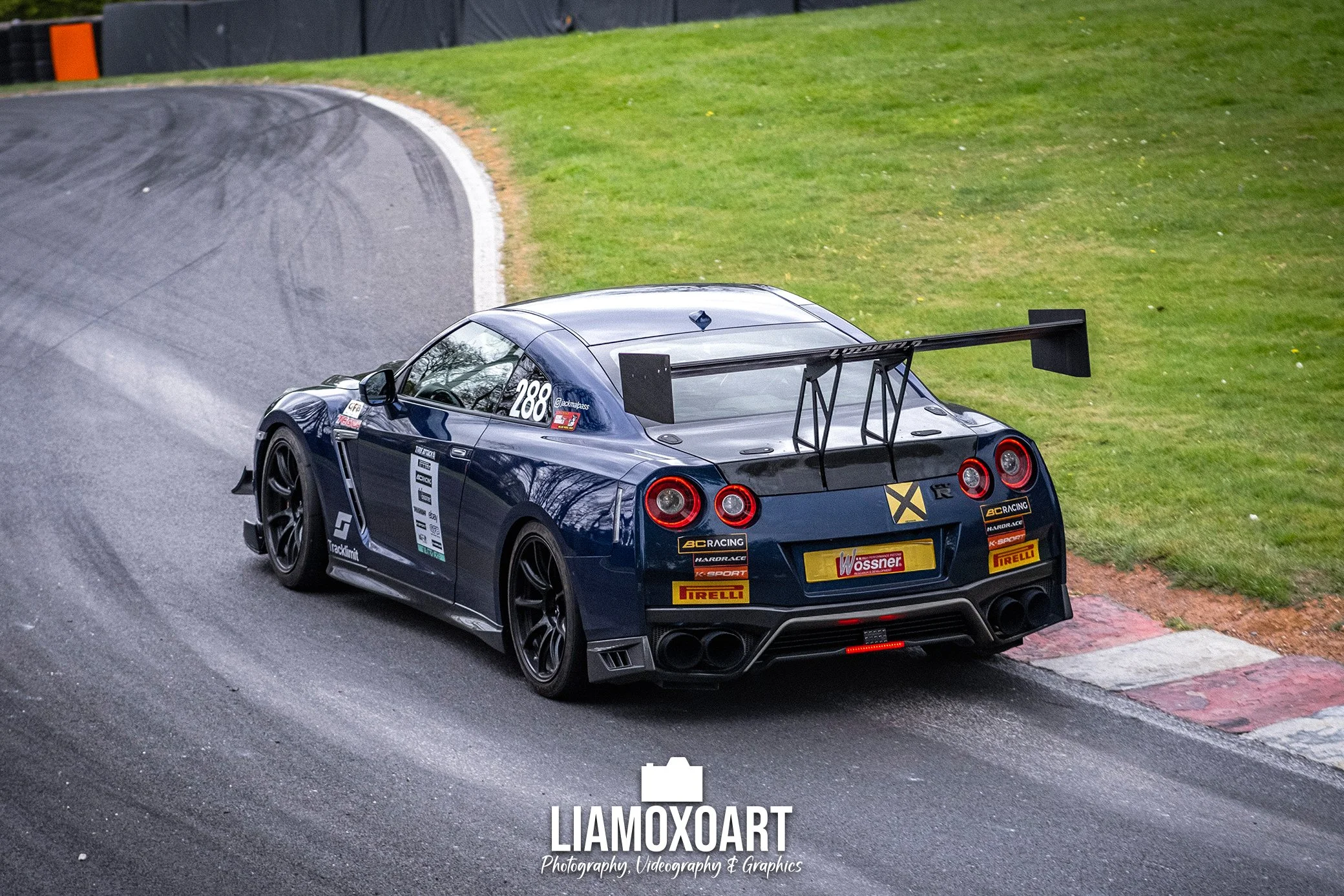 A race car on a race track, taking a turn on the asphalt with grass beside it. The car is black with various sponsor decals, large rear wing, and racing tires.
