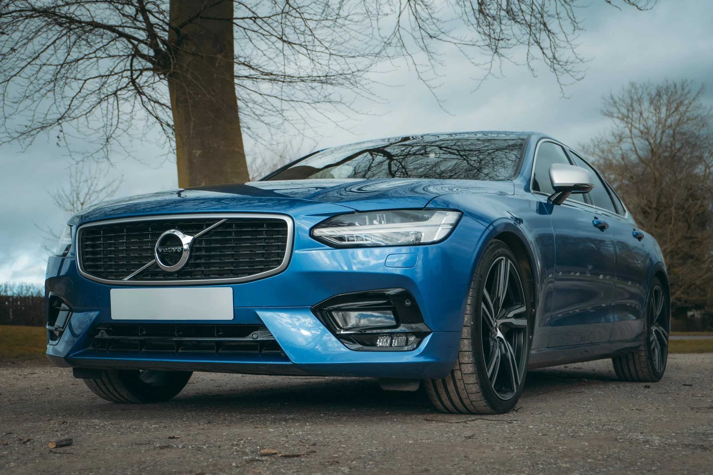 Blue Volvo sedan parked outdoors on a dirt surface with leafless trees and cloudy sky in the background.