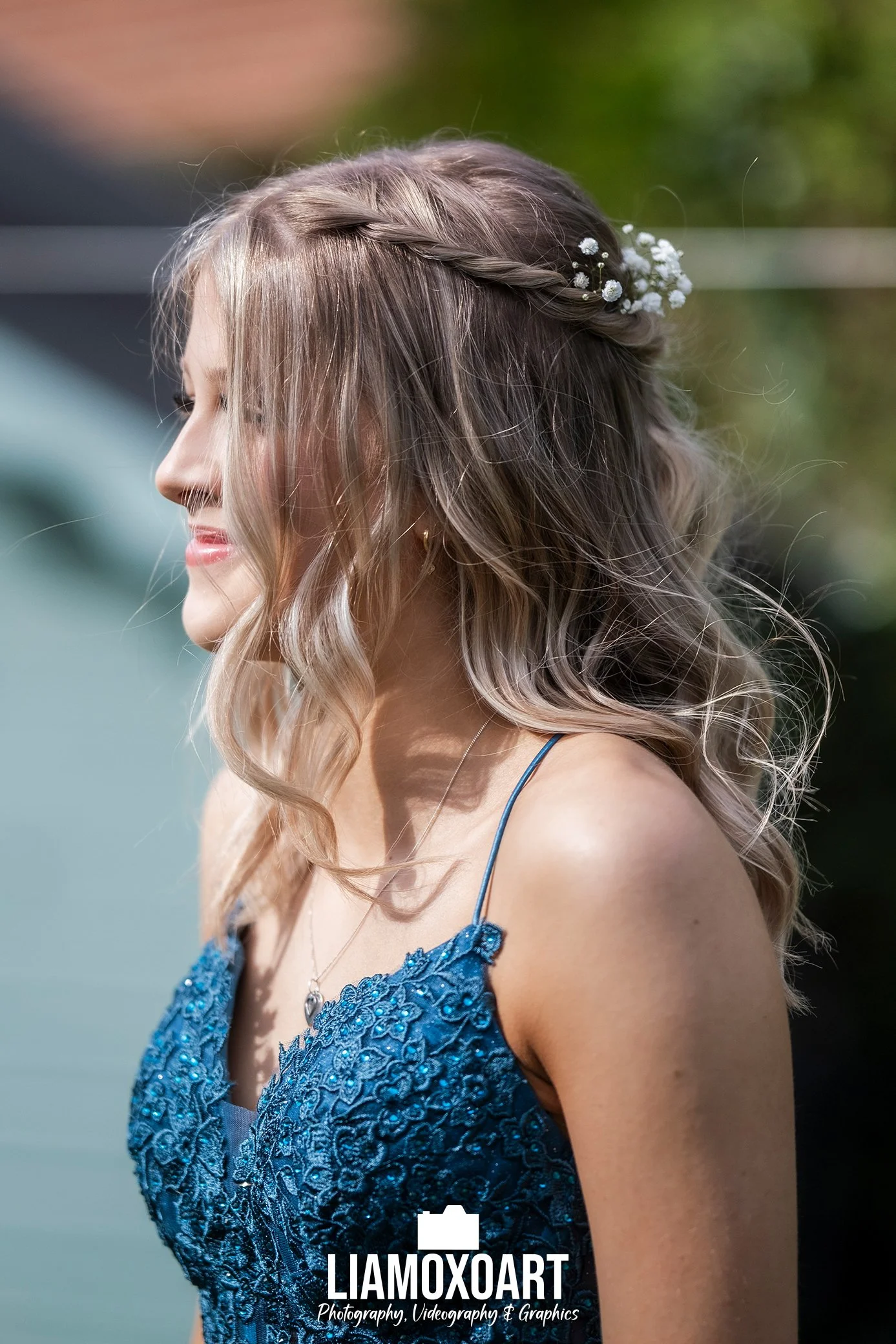 Side profile of a woman with wavy hair styled with a braid and small white flowers, wearing a blue lace dress with thin straps and a small pendant necklace, outdoors with green and orange blurred background.