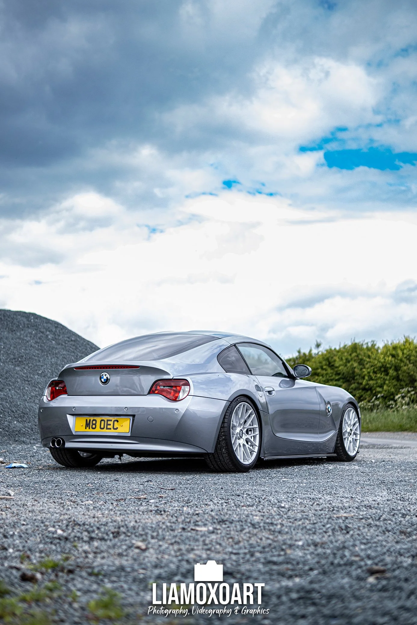 A silver BMW car parked on a gravel surface under a cloudy sky, with a background of greenery and a large pile of black gravel or rocks.