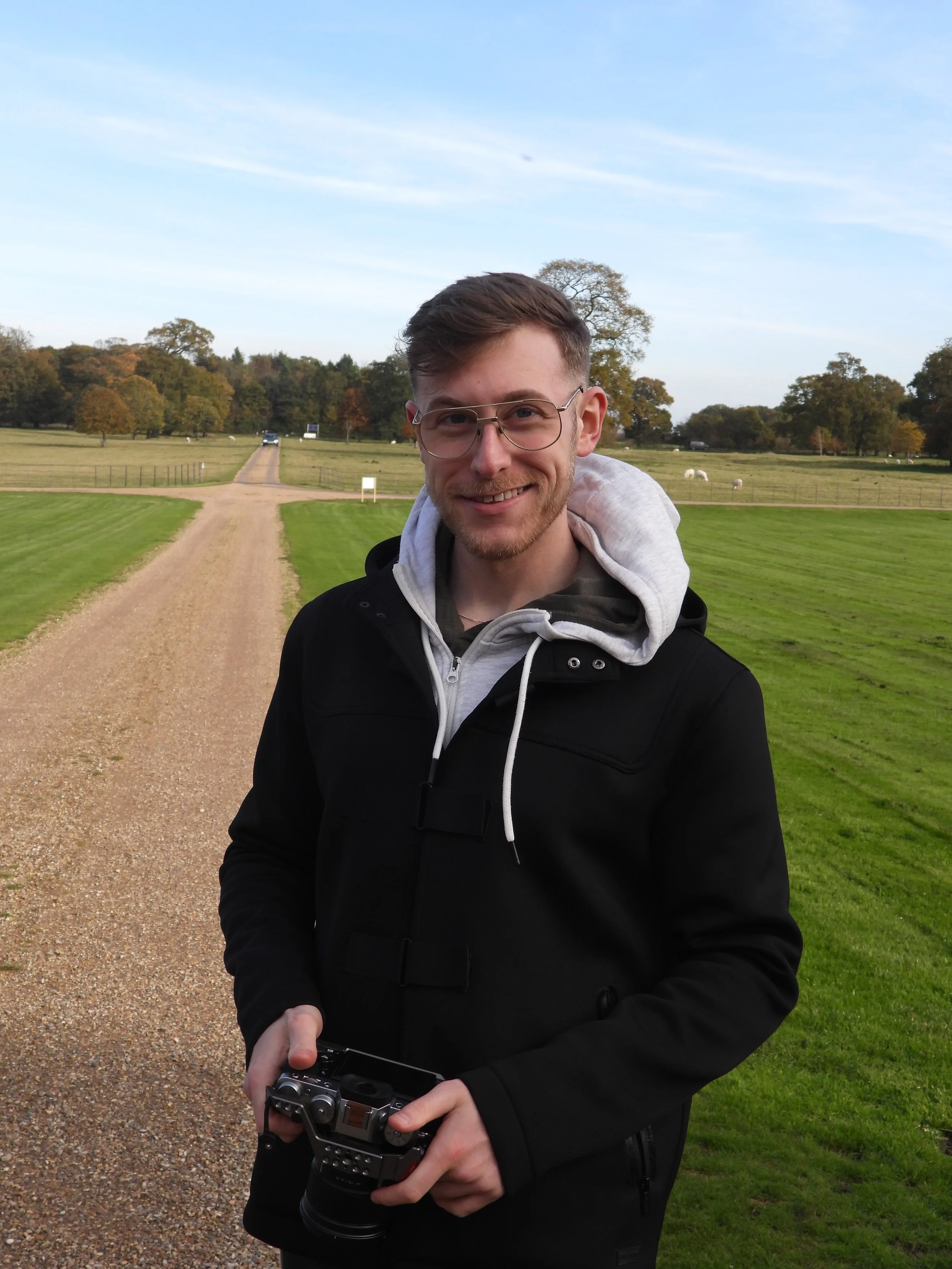 Young man with glasses holding a camera, standing on a dirt path in a grassy field with trees and sheep in the background during daytime.