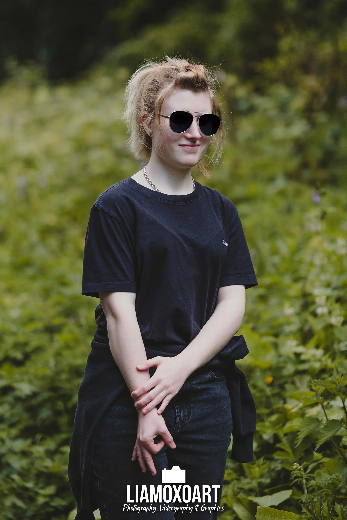 A young woman with short, wavy blonde hair wearing a black T-shirt and large black sunglasses, standing outdoors in front of green foliage, with a slightly smiley expression.