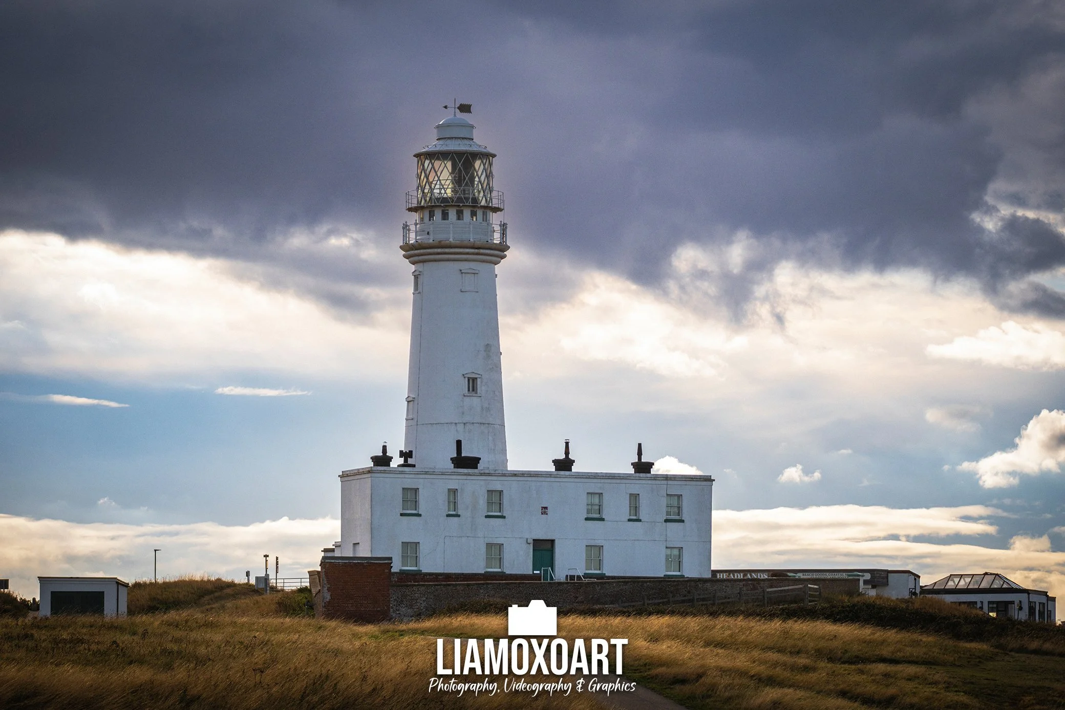 A tall white lighthouse with a black railing around its top, standing on a grassy area with a cloudy sky in the background.