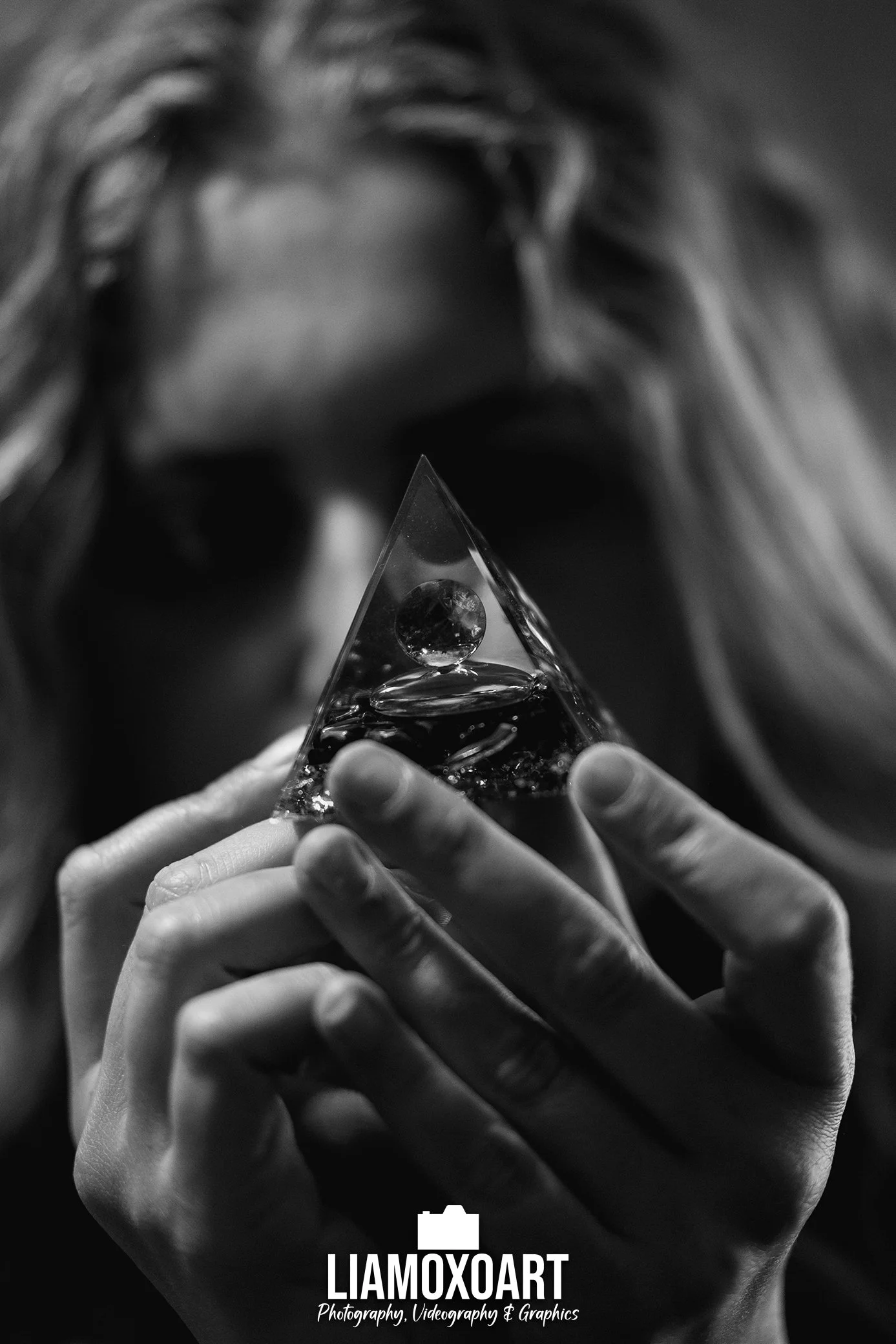 A black and white close-up photograph of a person holding a triangular glass object with a sphere inside it. The person's face is blurred in the background. There is a logo and text at the bottom of the image that reads 'LIAMOXOART Photography, Video