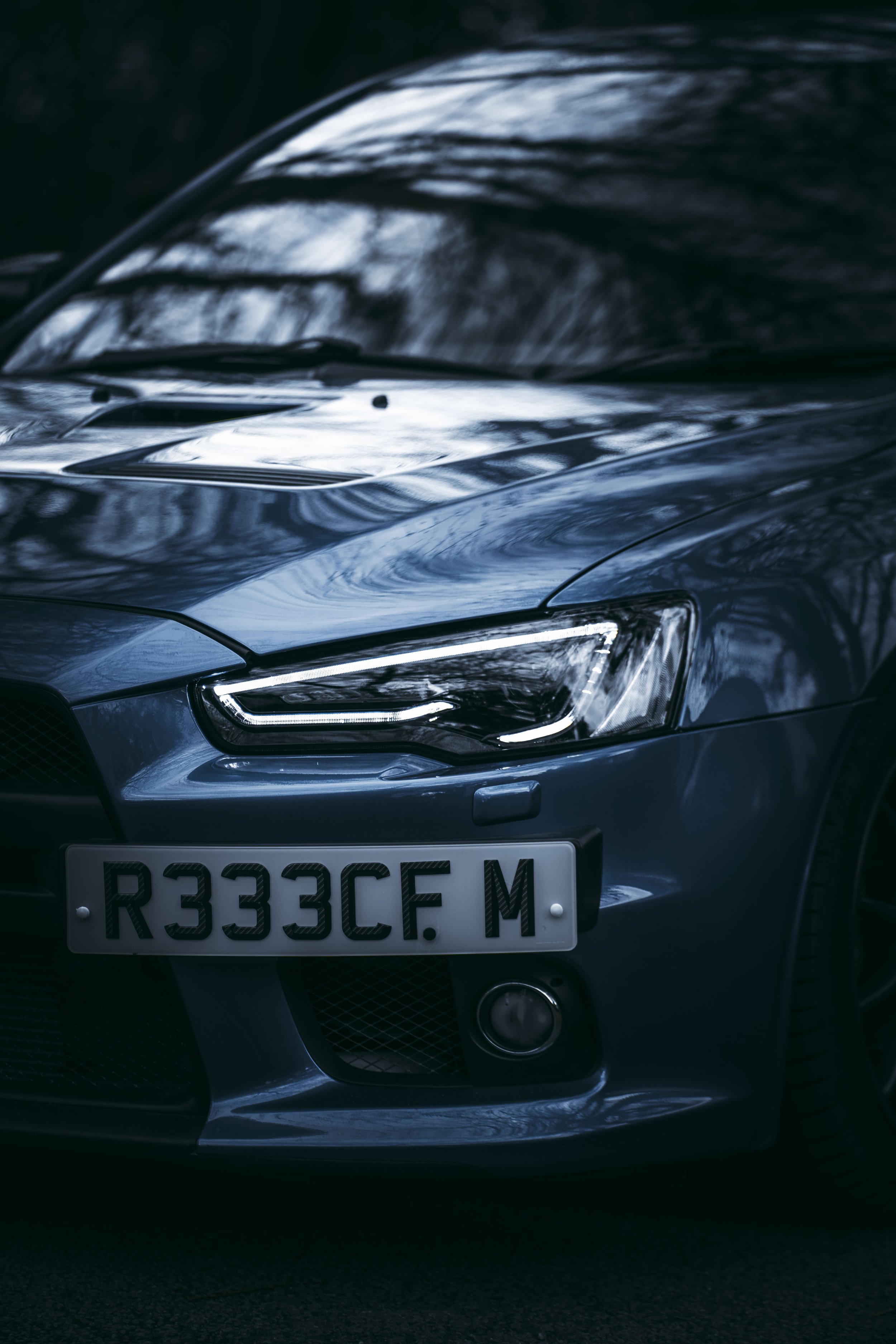Close-up of a dark blue car's front, showing headlight, part of the hood, and a custom license plate reading 'R333CF M'. Reflection of trees and sky are visible on the car's surface.