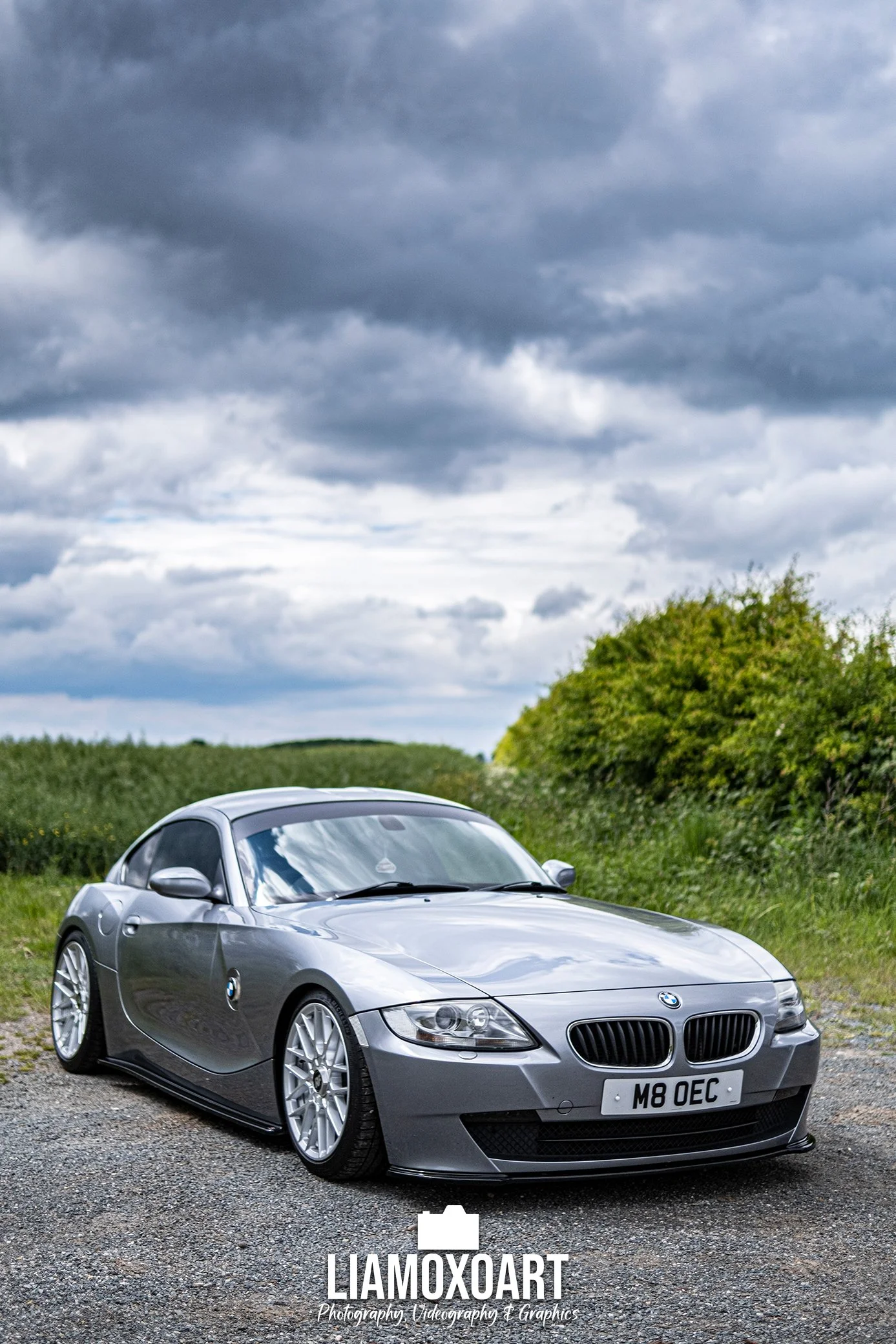 A silver BMW Z3 sports car parked on a gravel area with green bushes and trees in the background under a cloudy sky.