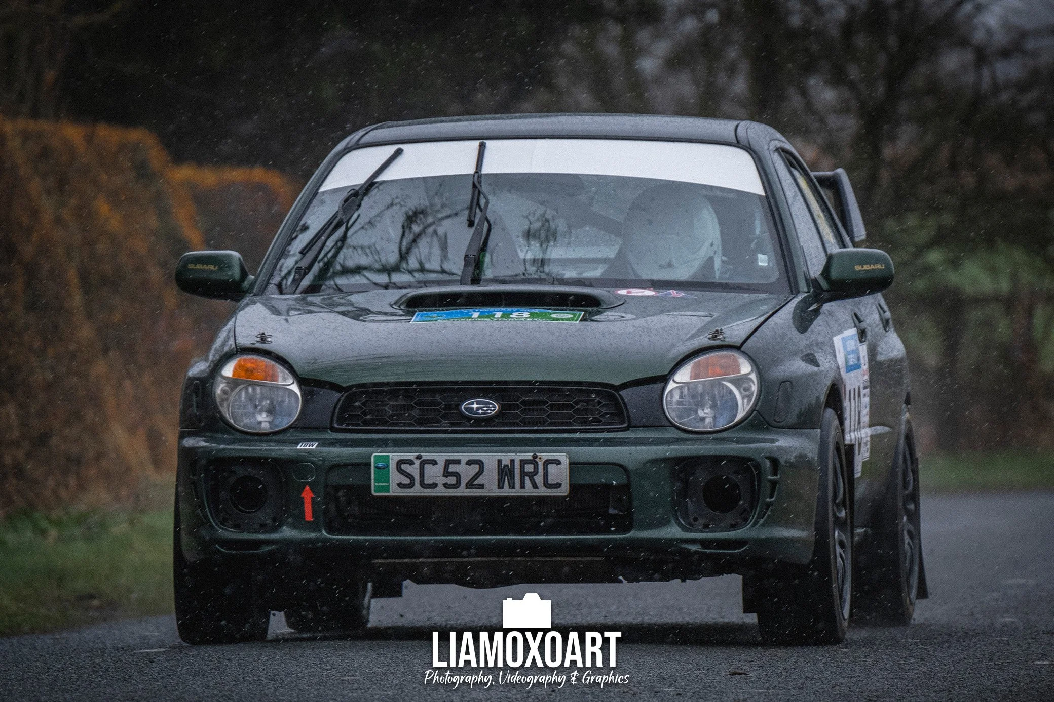 A black Subaru rally car with a partially obscured license plate, driving on a wet road in rainy weather, with rain droplets visible on the windshield and trees in the background.